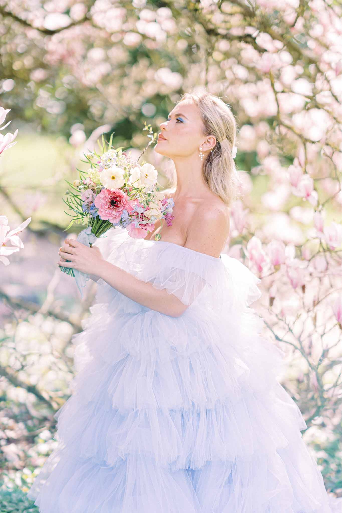 Bride in pale blue tulle gown with mixed bouquet posing among flowering shrubs in garden setting