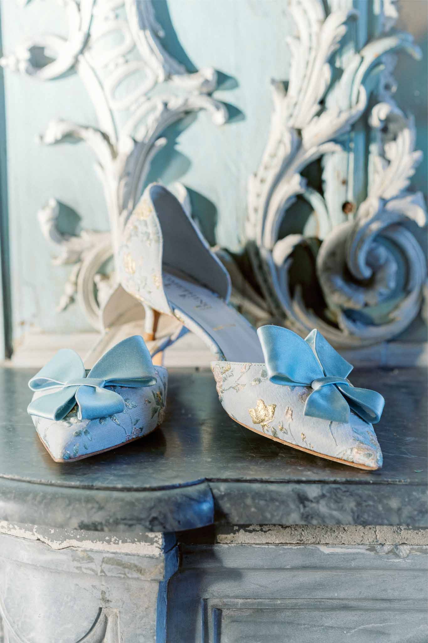 Bride's ivory and gold flat shoes with blue silk bows on ornate stone ledge at classical venue