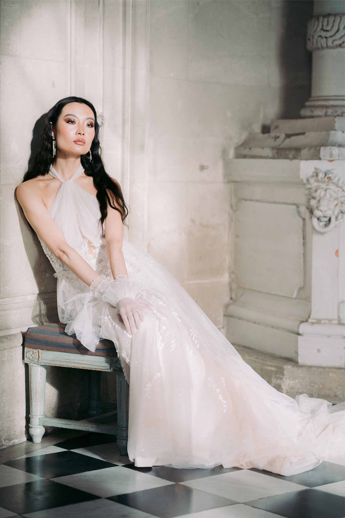 Bride in ivory halter dress seated on vintage chair in neoclassical interior with checkered floor
