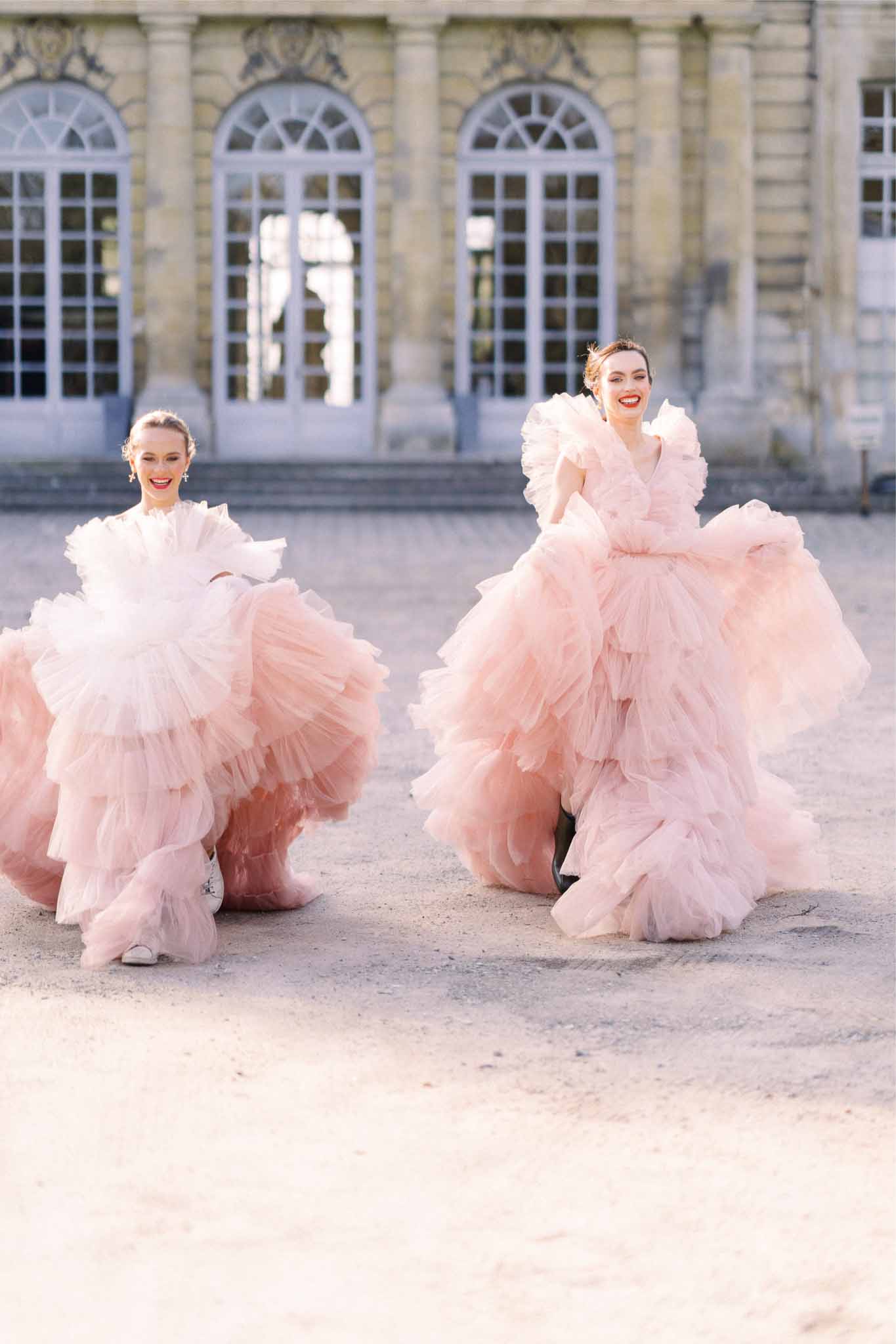 Two women in blush tulle gowns posed in classical stone courtyard with arched windows