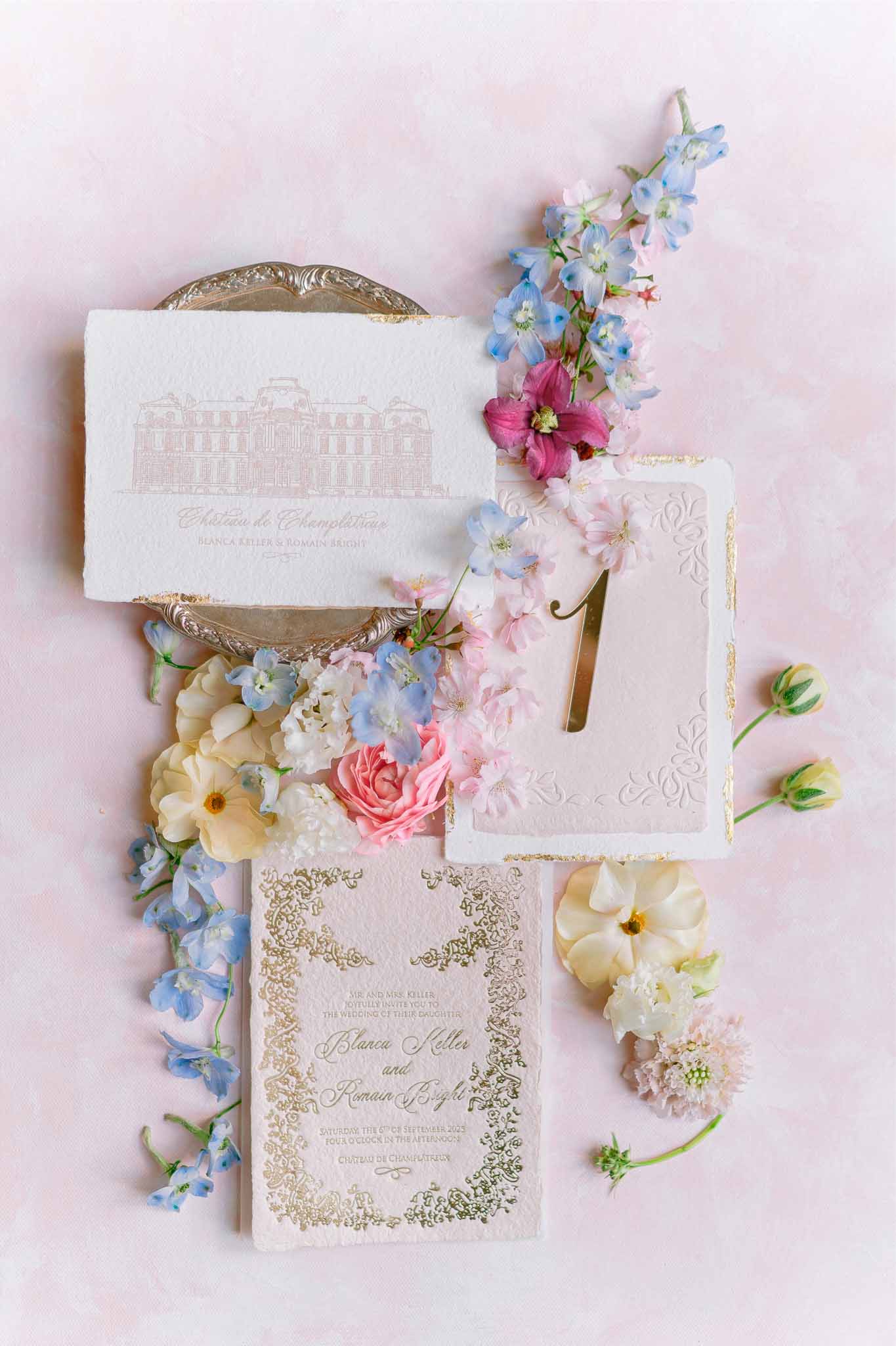Wedding stationery flat lay with cream invitations, gold details, and pastel flowers on pink background