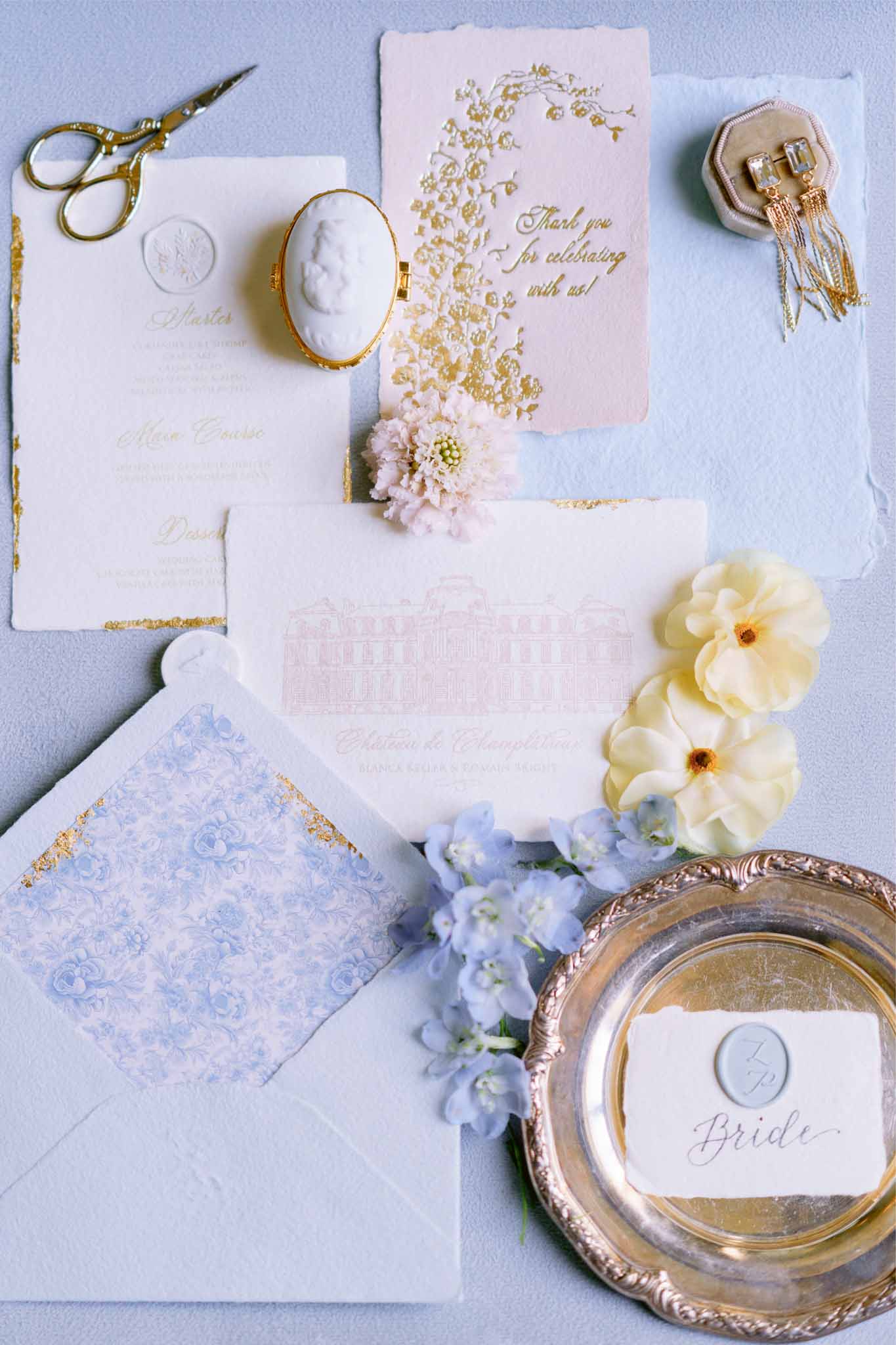 Wedding invitation flat lay with cream stationery, blush flowers, and gold accessories on light blue surface