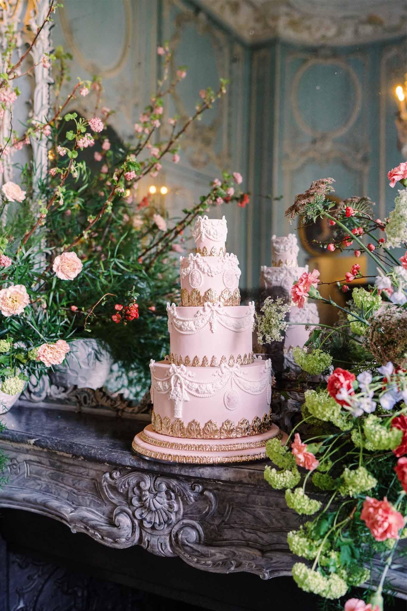 Four-tier wedding cake with blush fondant and gold accents surrounded by coral and pink floral arrangements