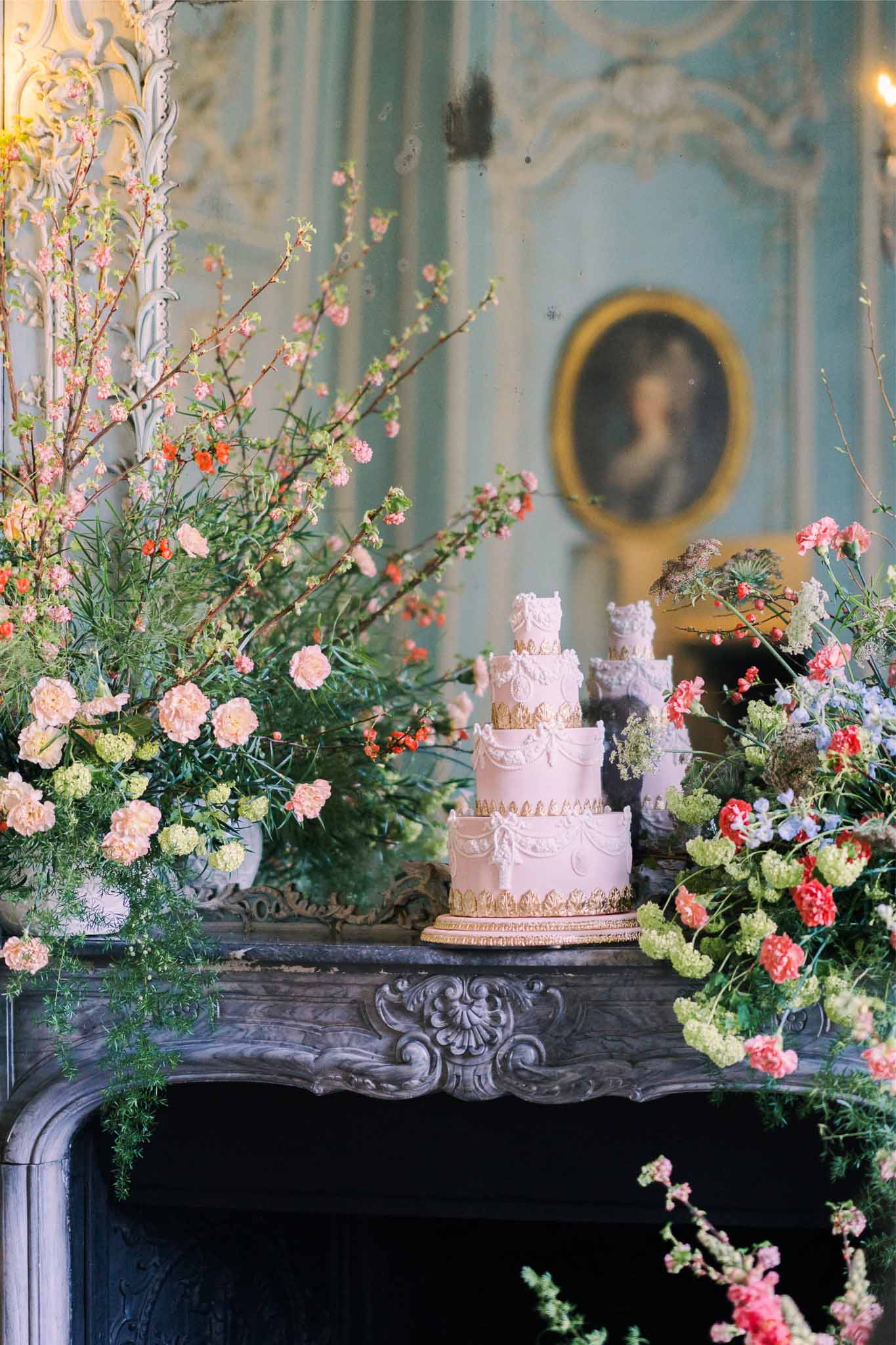 Five-tiered blush wedding cake with floral arrangements in classical interior with ornate fireplace mantel