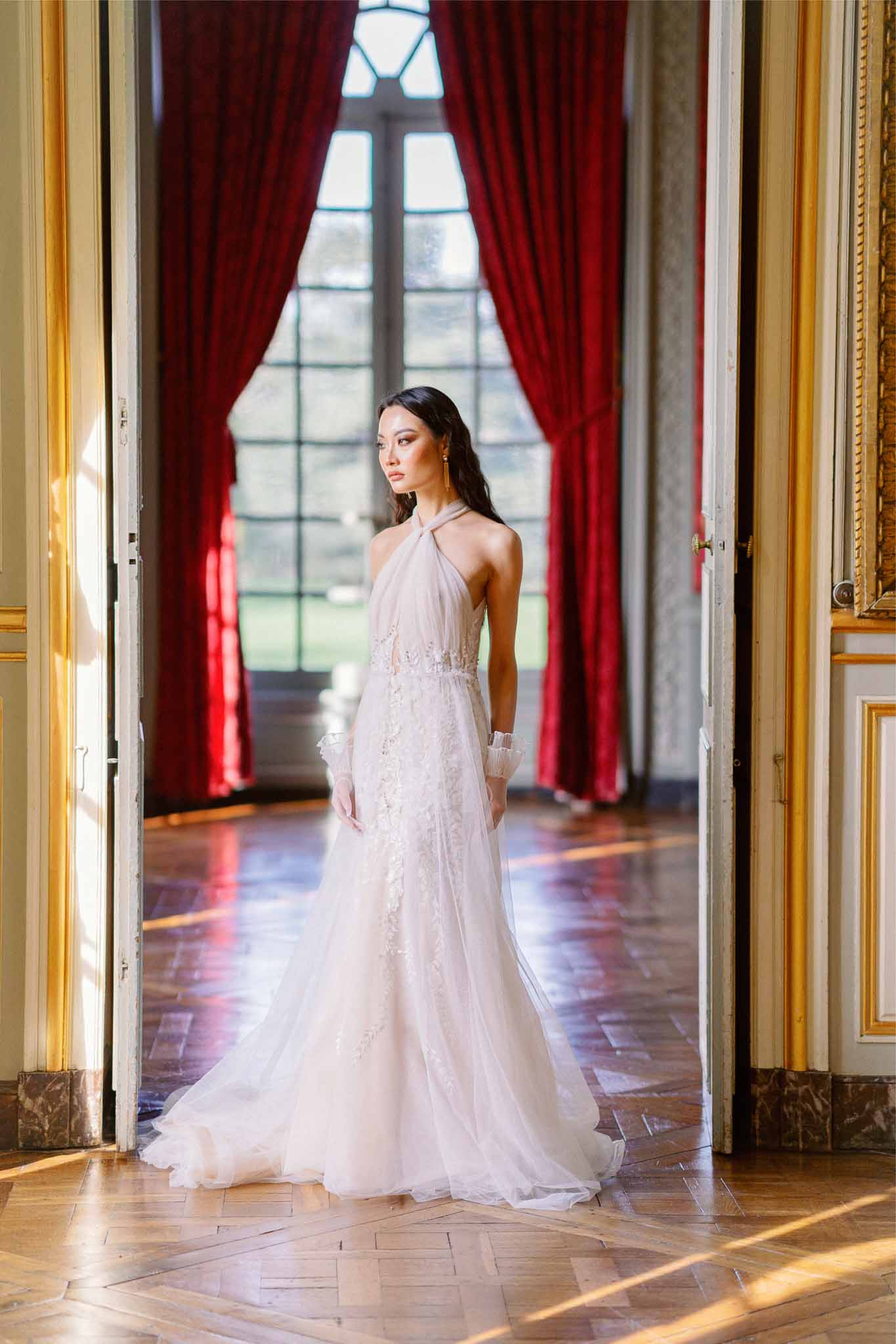 Bride in blush wedding gown standing in classical interior hallway with gilt trim and burgundy curtains