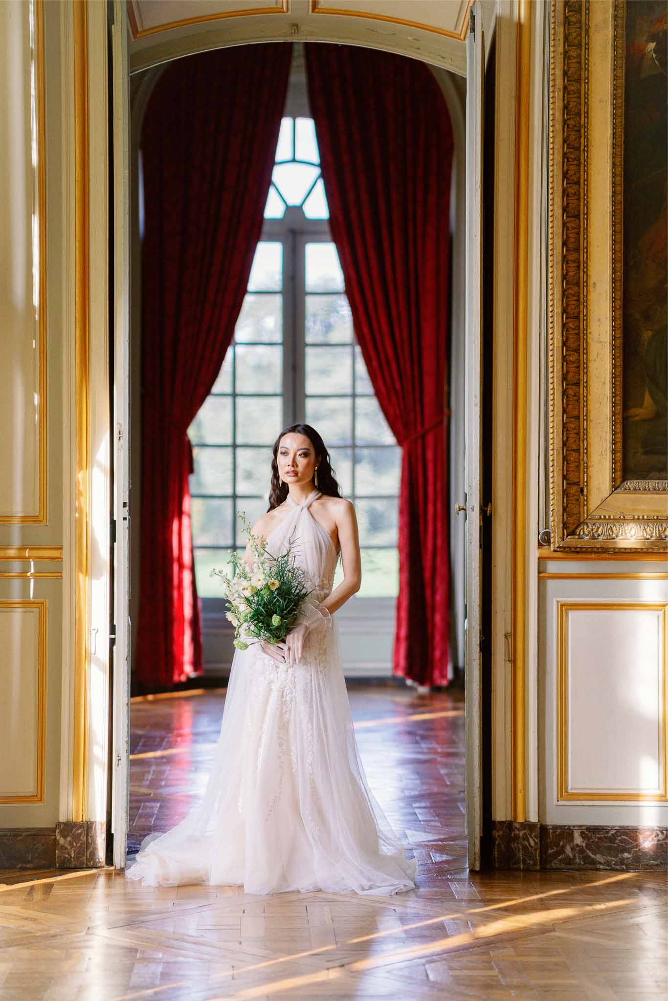 Bride in blush gown with bouquet posing in formal palace corridor with classical architecture
