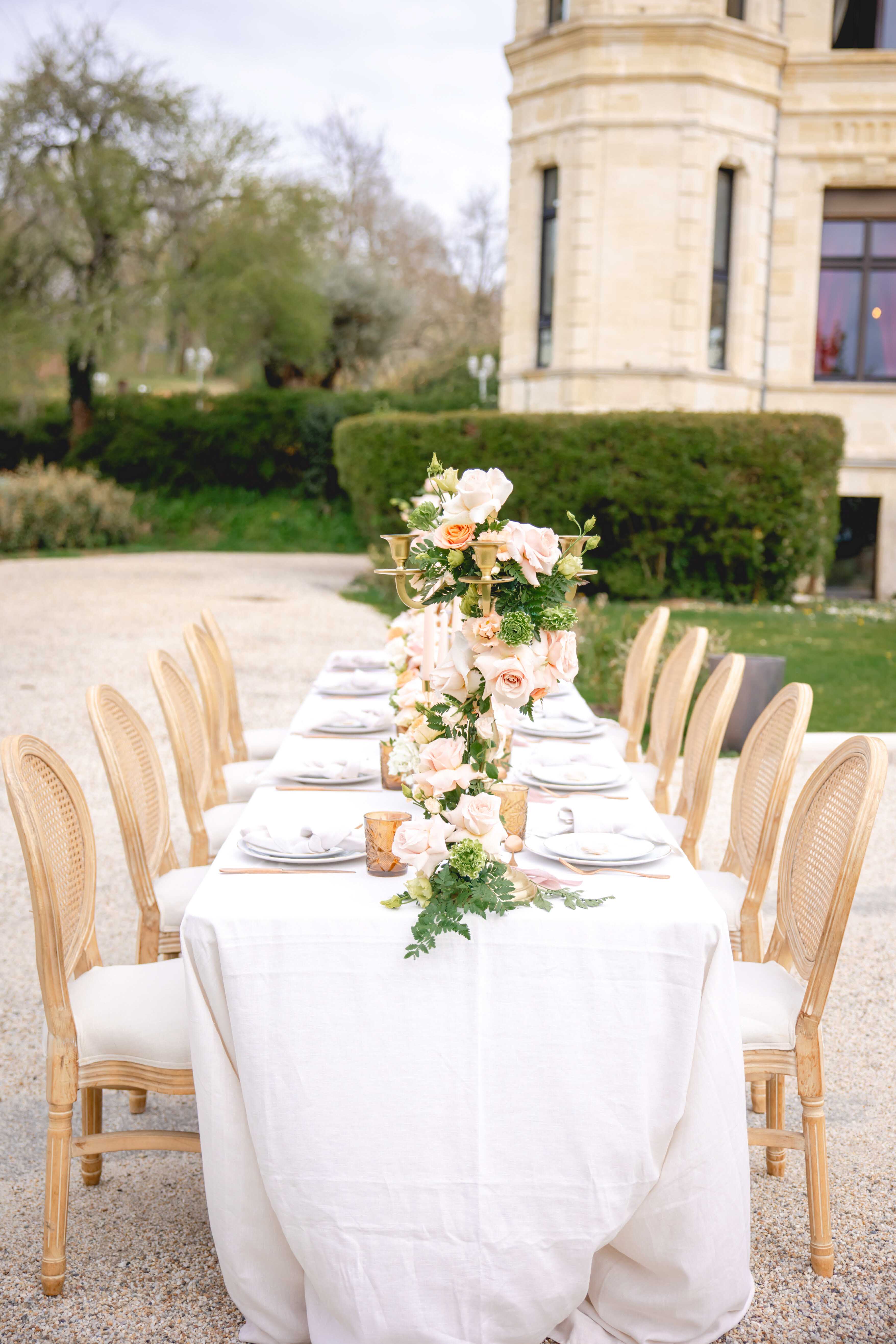 Long reception table with gold candelabra, blush and peach floral runner, and cane-back chairs on chateau courtyard