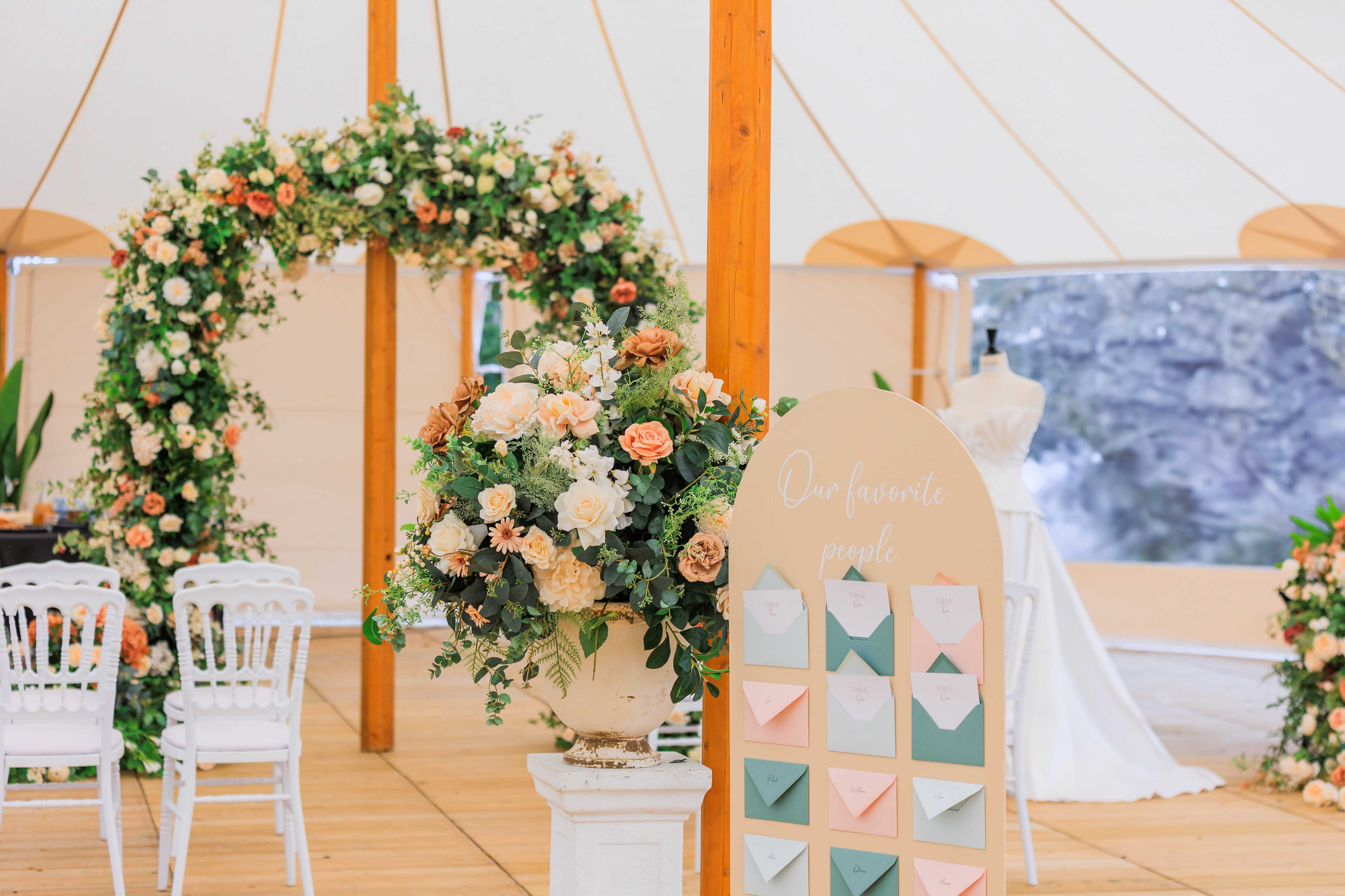 Decorated sperry tent with seating chart sign, peach and cream floral urn, and circular flower arch for ceremony