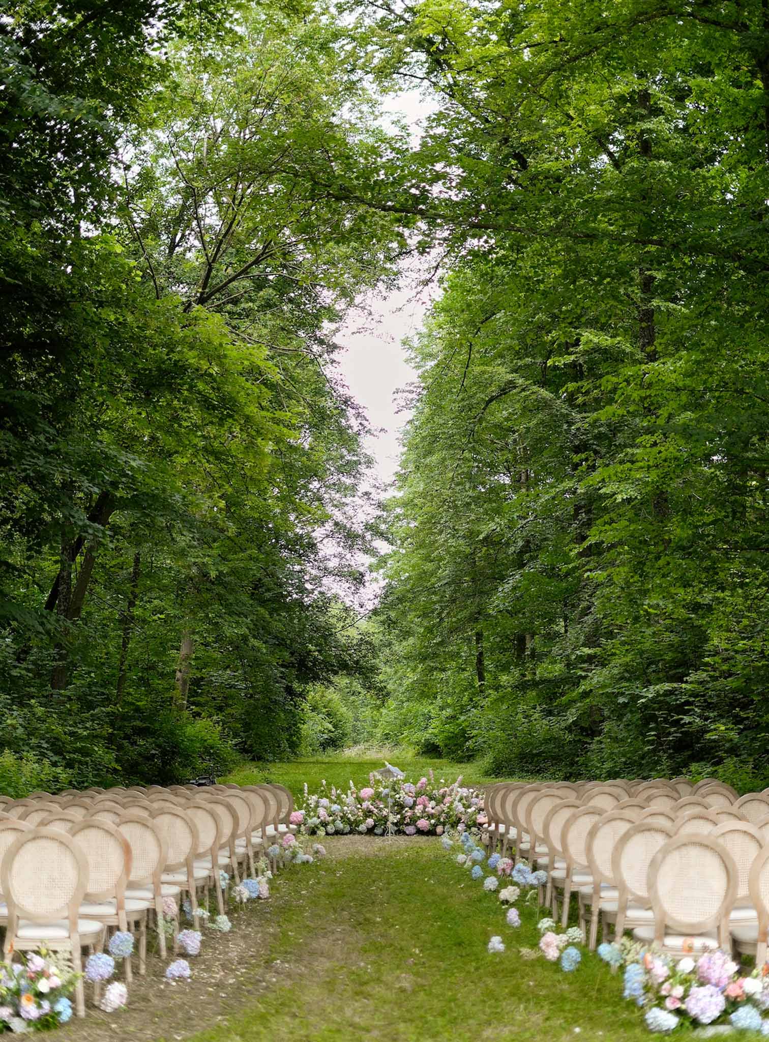 Outdoor wedding ceremony setup with cream chairs and hydrangea aisle in forest clearing