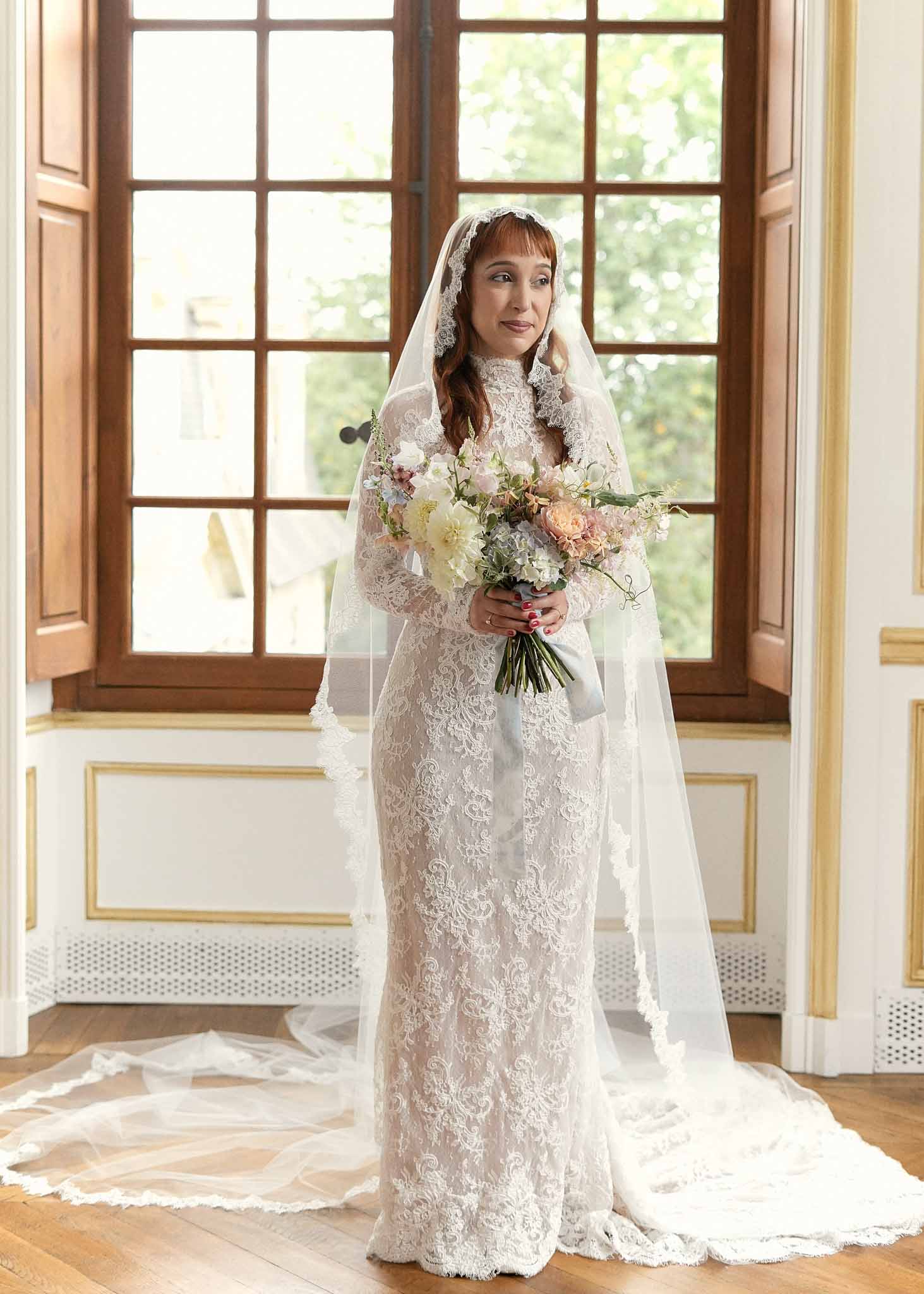 Bride in ivory lace wedding dress holding bouquet in elegant interior hallway with gold accents
