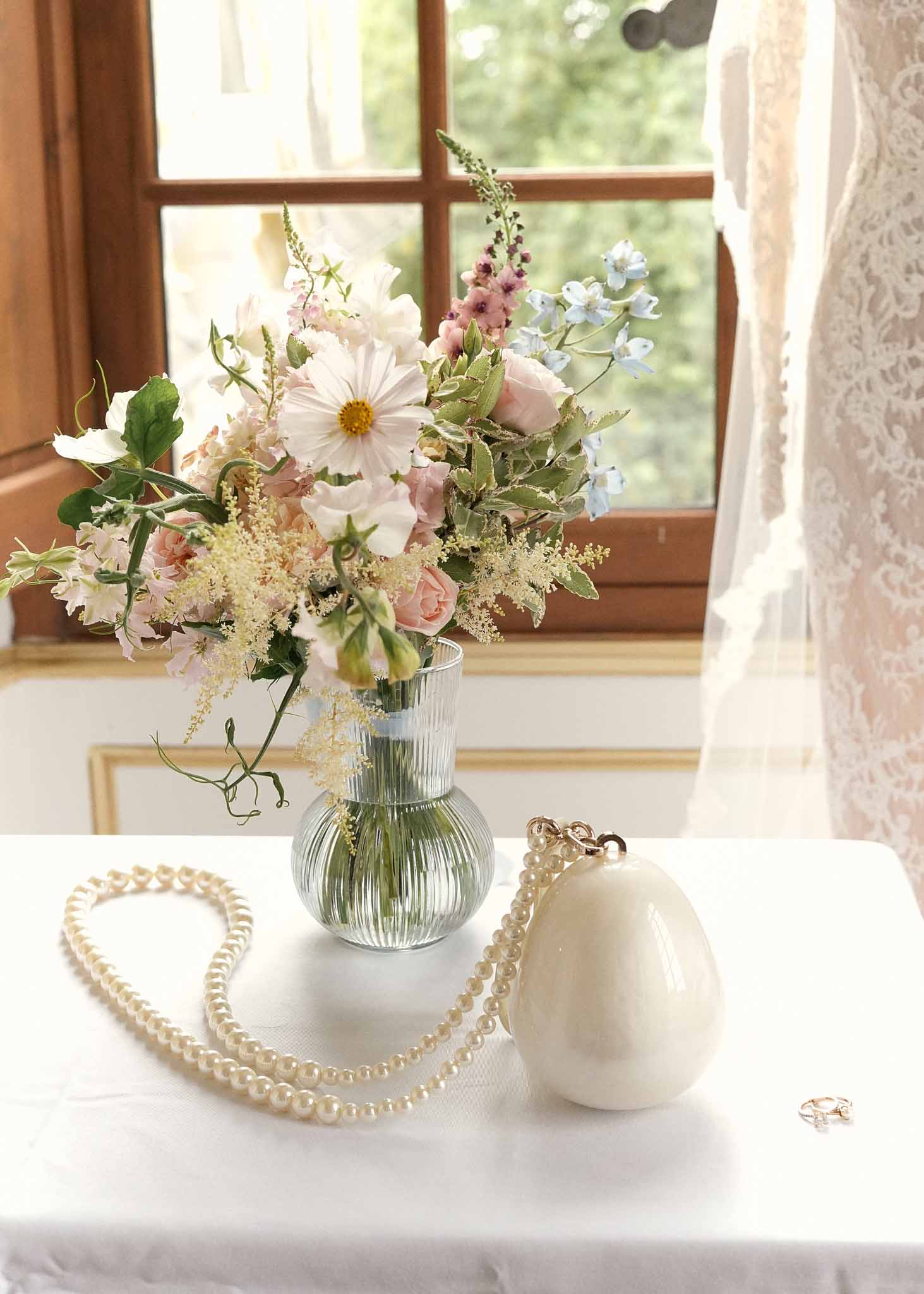 Bridal accessories and floral arrangement flat lay on white linen beside window