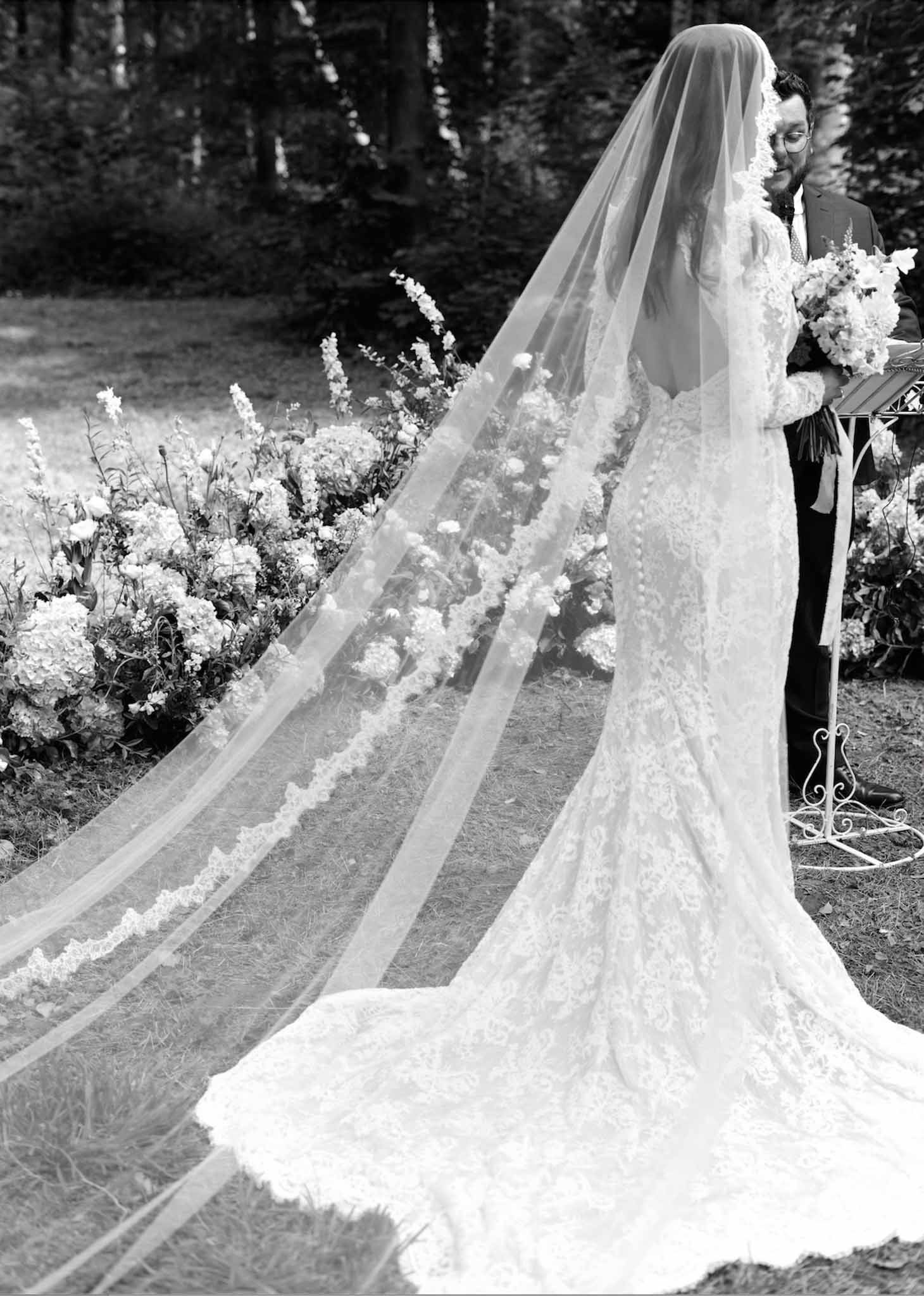 Bride and groom during outdoor wedding ceremony in wooded garden with cathedral veil and white flowers