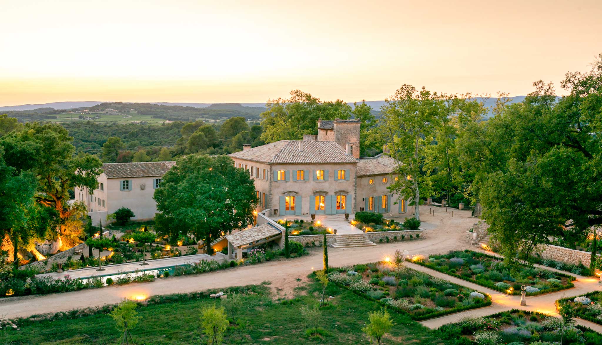 Aerial dusk view of Provencal stone bastide with swimming pool, parterre garden, and glowing interior lights