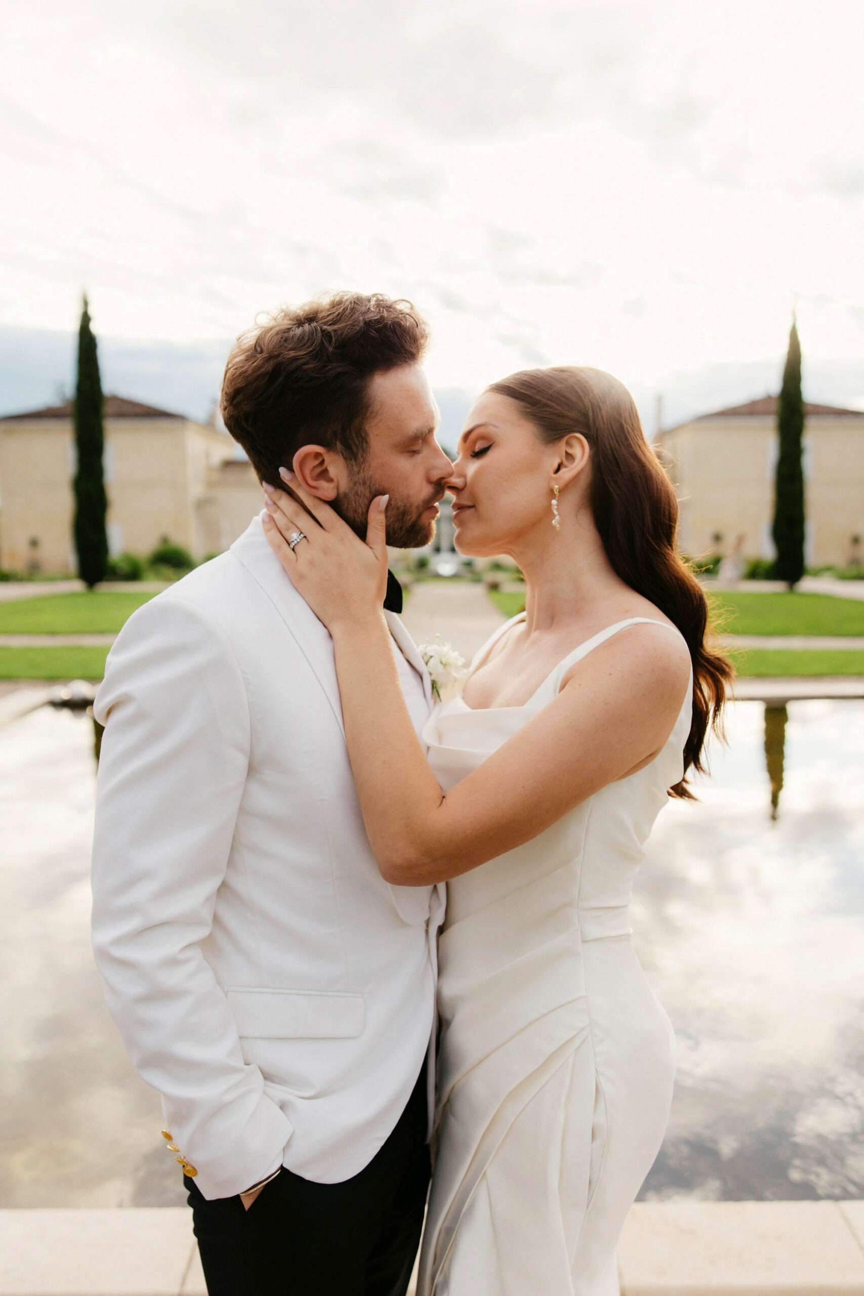 Bride in ivory slip gown and groom in white tuxedo kissing by reflecting pool at formal chateau garden
