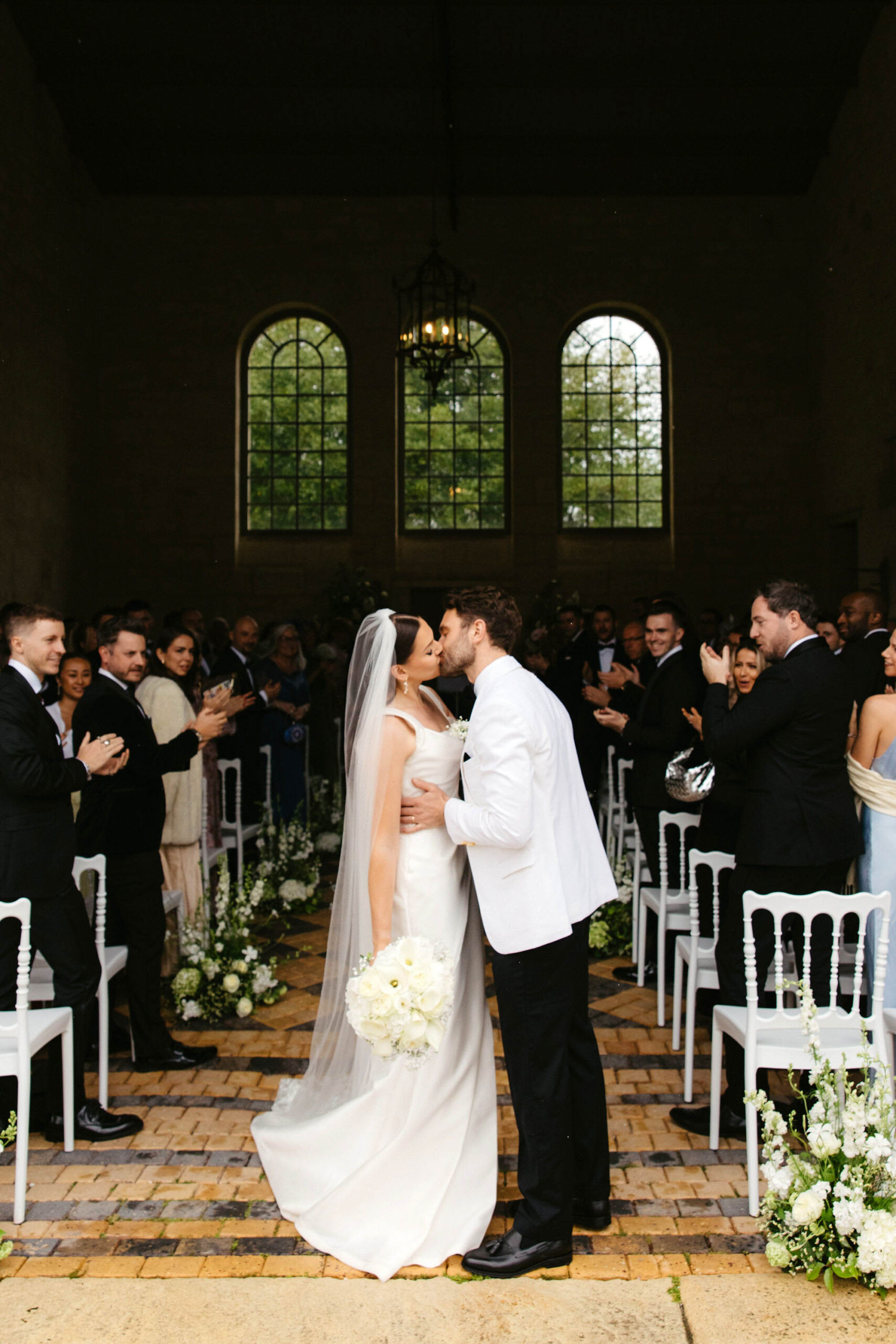 Bride and groom share first kiss in stone chapel with arched windows and white hydrangea aisle arrangements