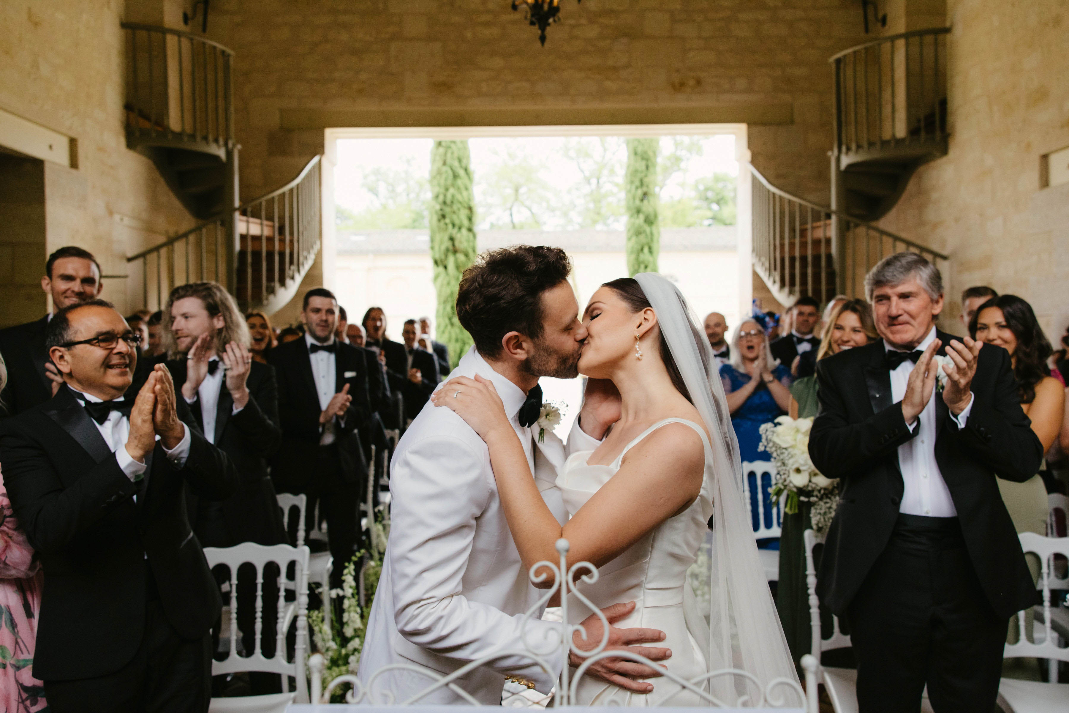 Bride and groom first kiss as married couple in stone chapel with guests applauding