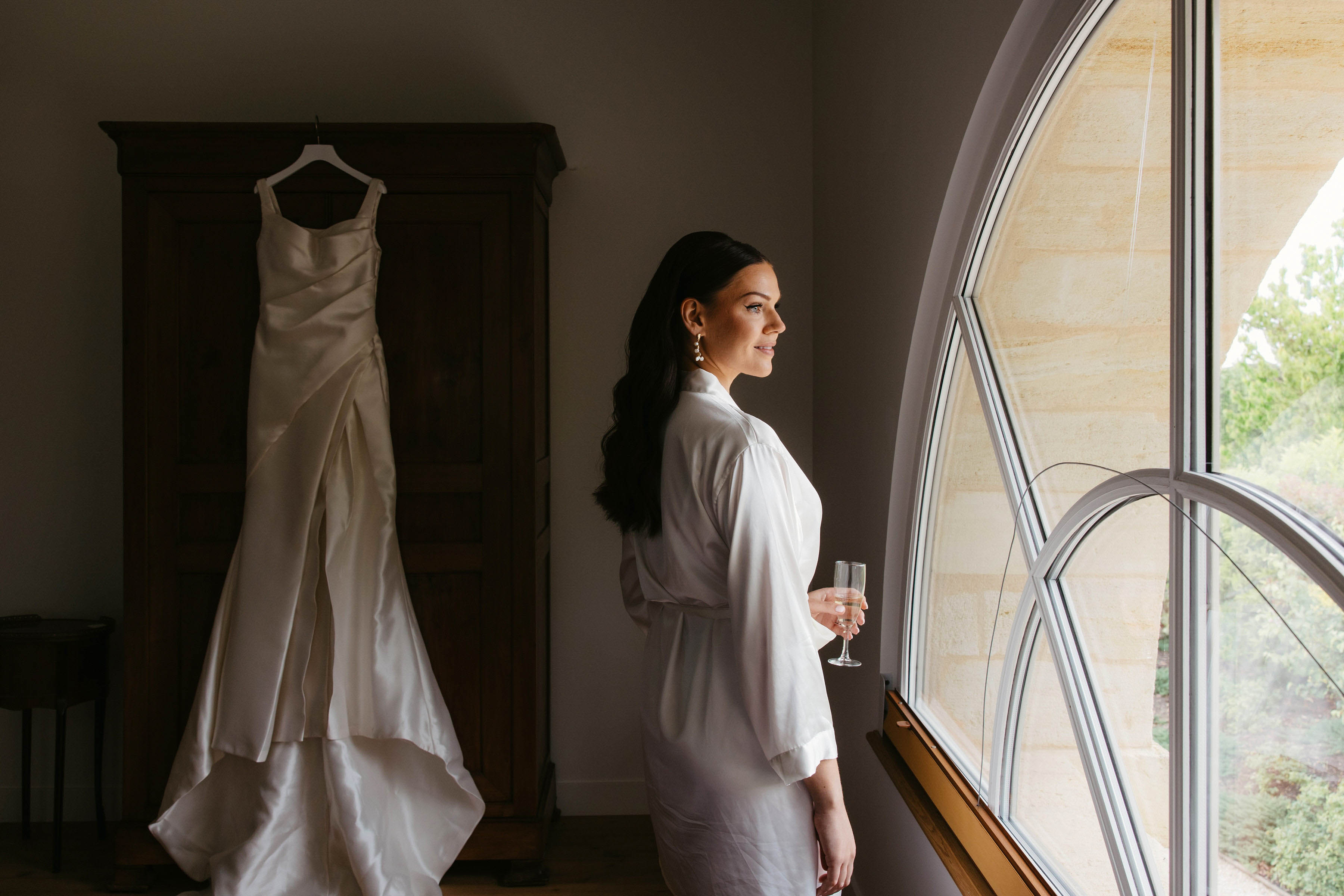 Bride in silk robe holding champagne flute by window in preparation room with wedding dress hanging in background