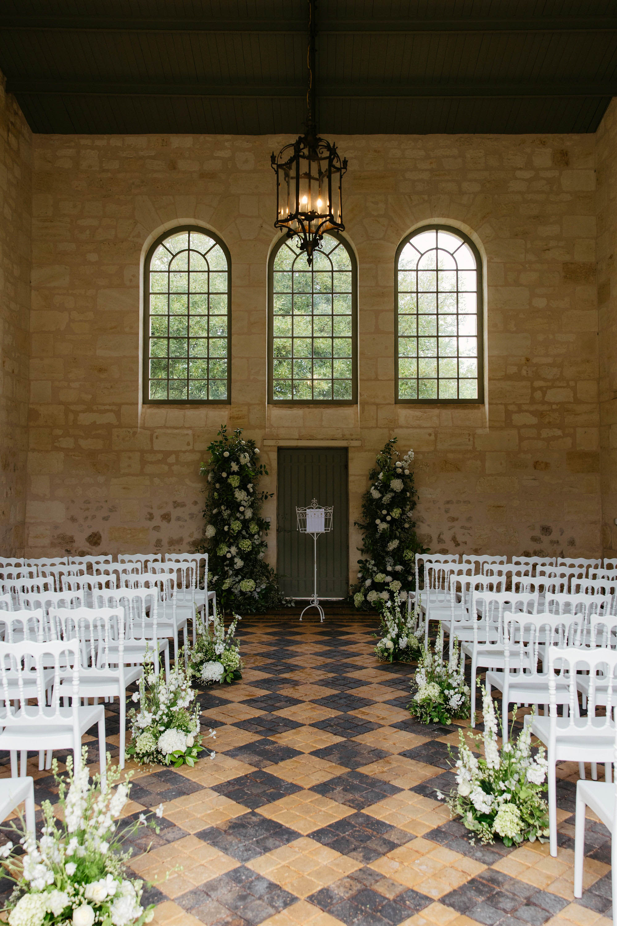 Eighty Napoleon chairs facing white floral columns in limestone chapel with arched windows and iron chandelier