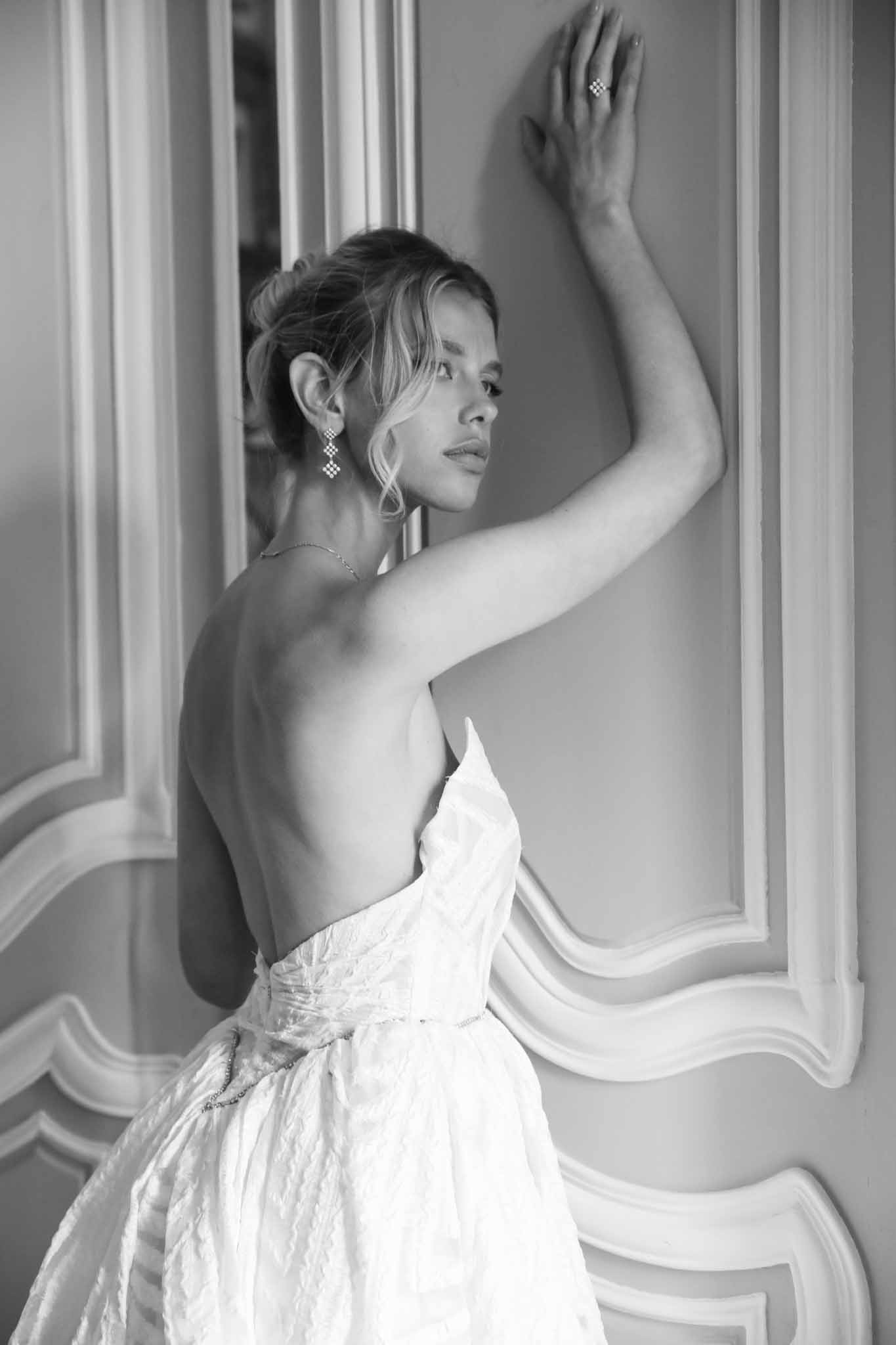 Black and white portrait of bride showing open-back strapless gown and crystal earrings against ornate wall panelling
