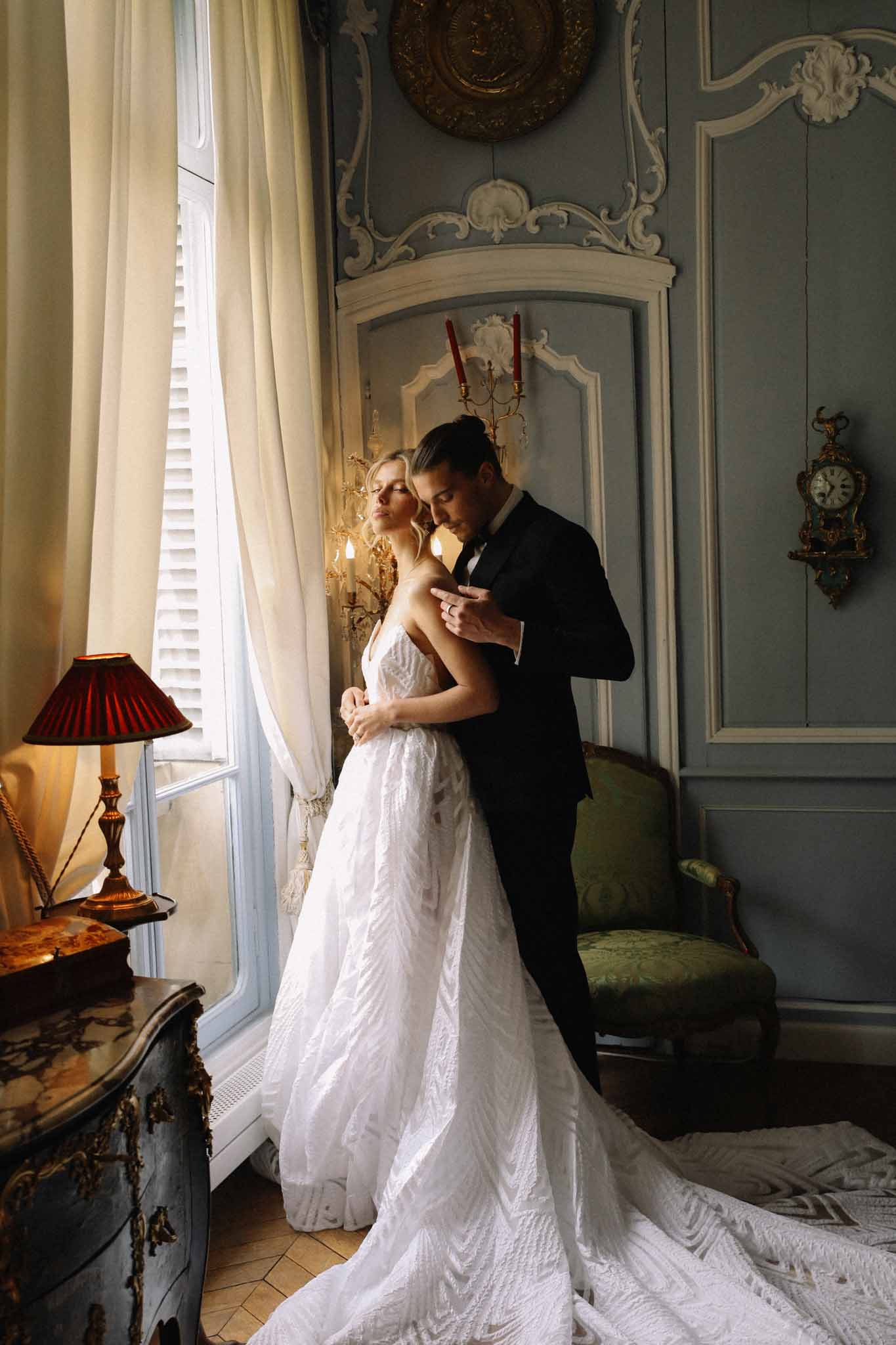 Bride in white lace gown and groom in black tuxedo embracing by window in blue-grey panelled chateau salon