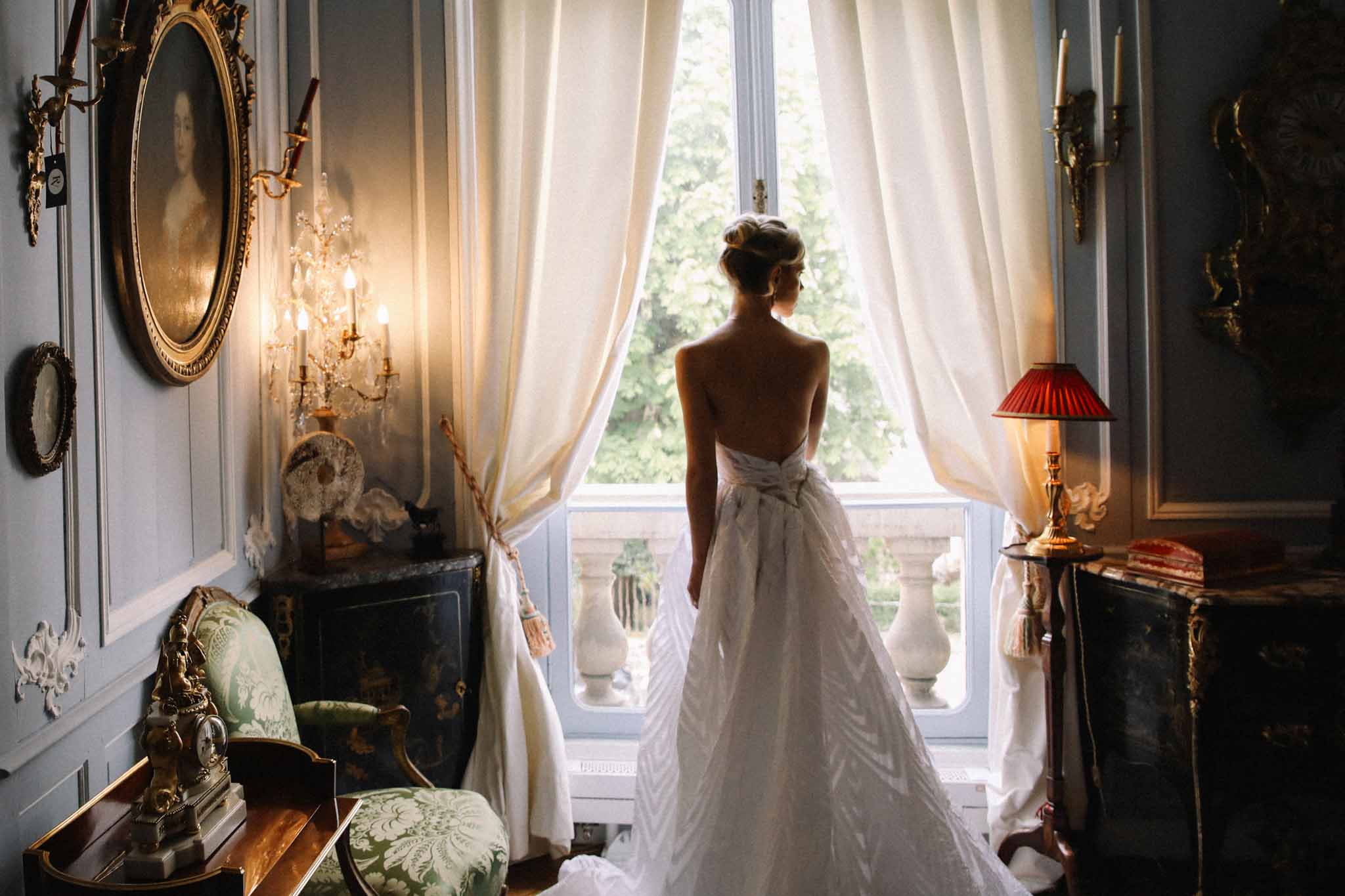 Bride from behind in bow-back ballgown at chateau window with blue-grey paneling and gilt portraits