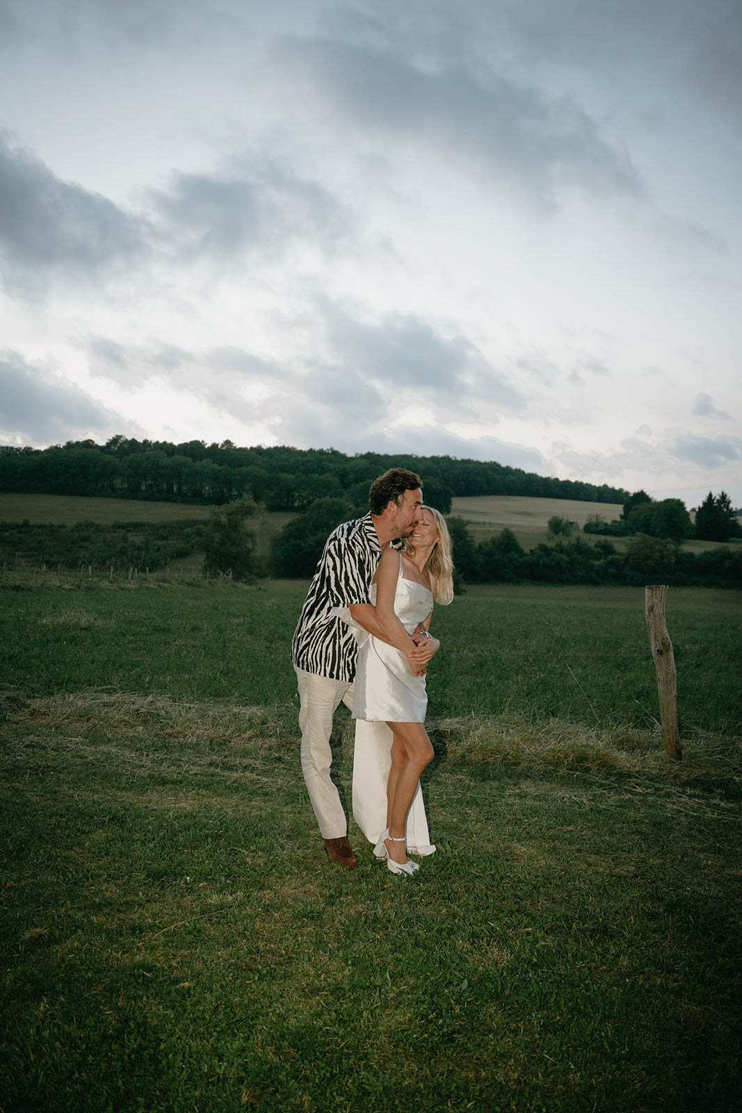 Couple embracing and kissing in rural field with rolling hills and forest background