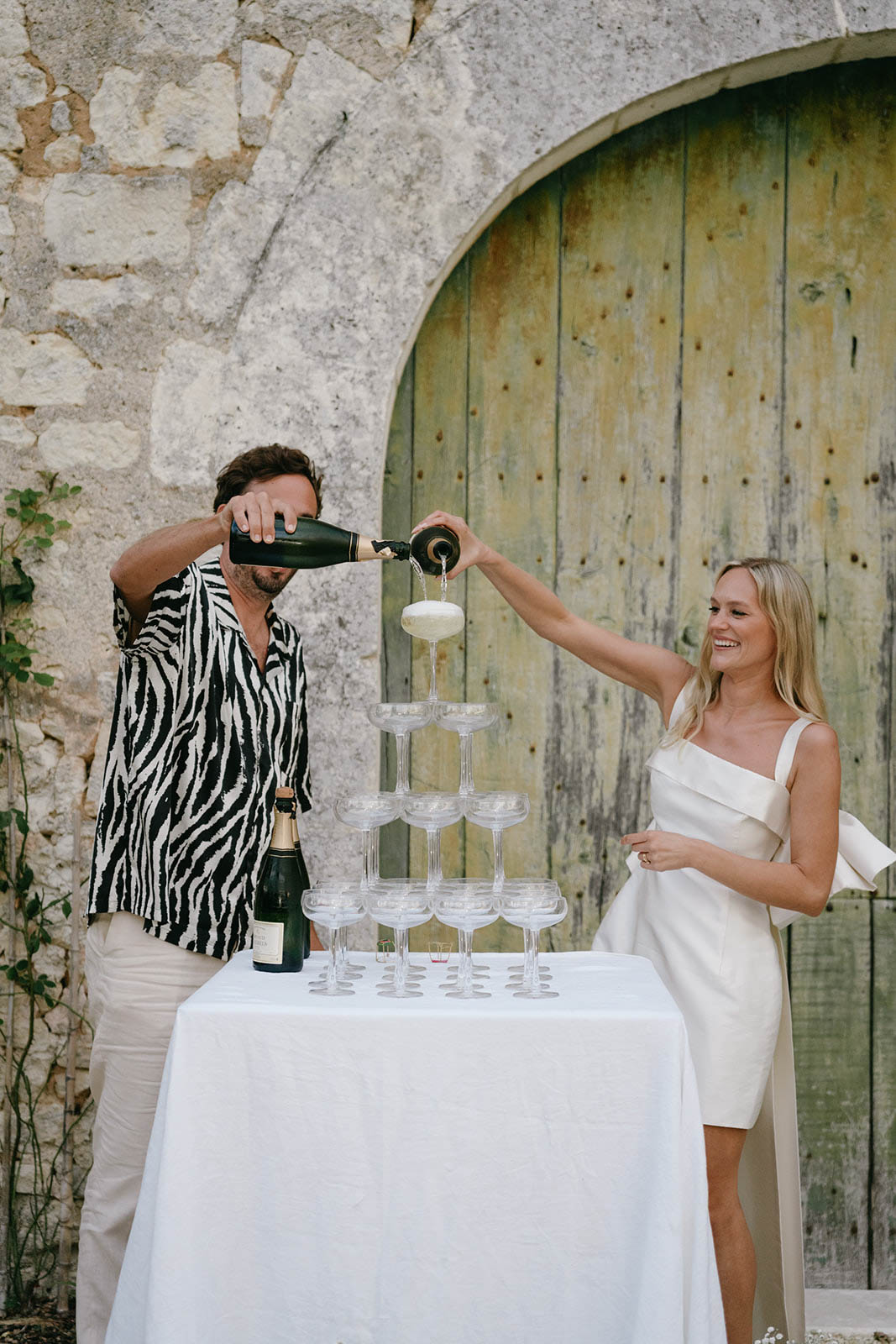 Couple performing champagne tower pour during outdoor reception at rustic stone venue
