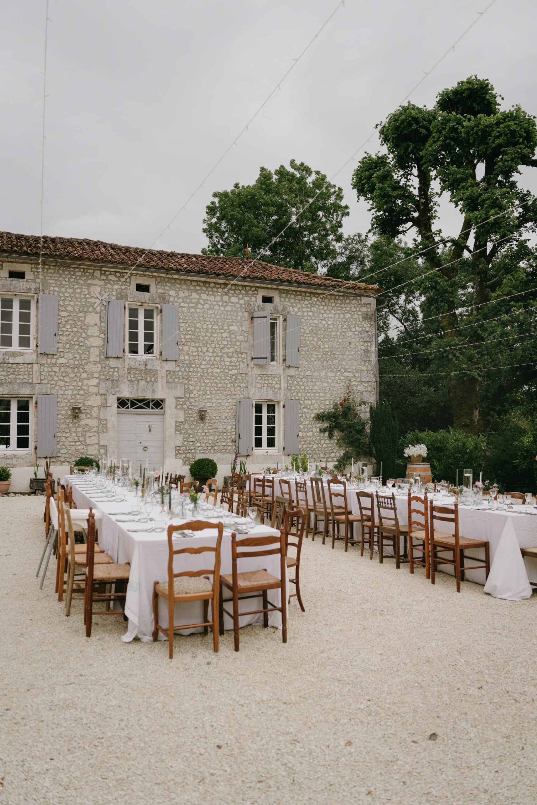 Outdoor courtyard reception with parallel banquet tables, rush-seat chairs, and fairy lights at stone farmhouse