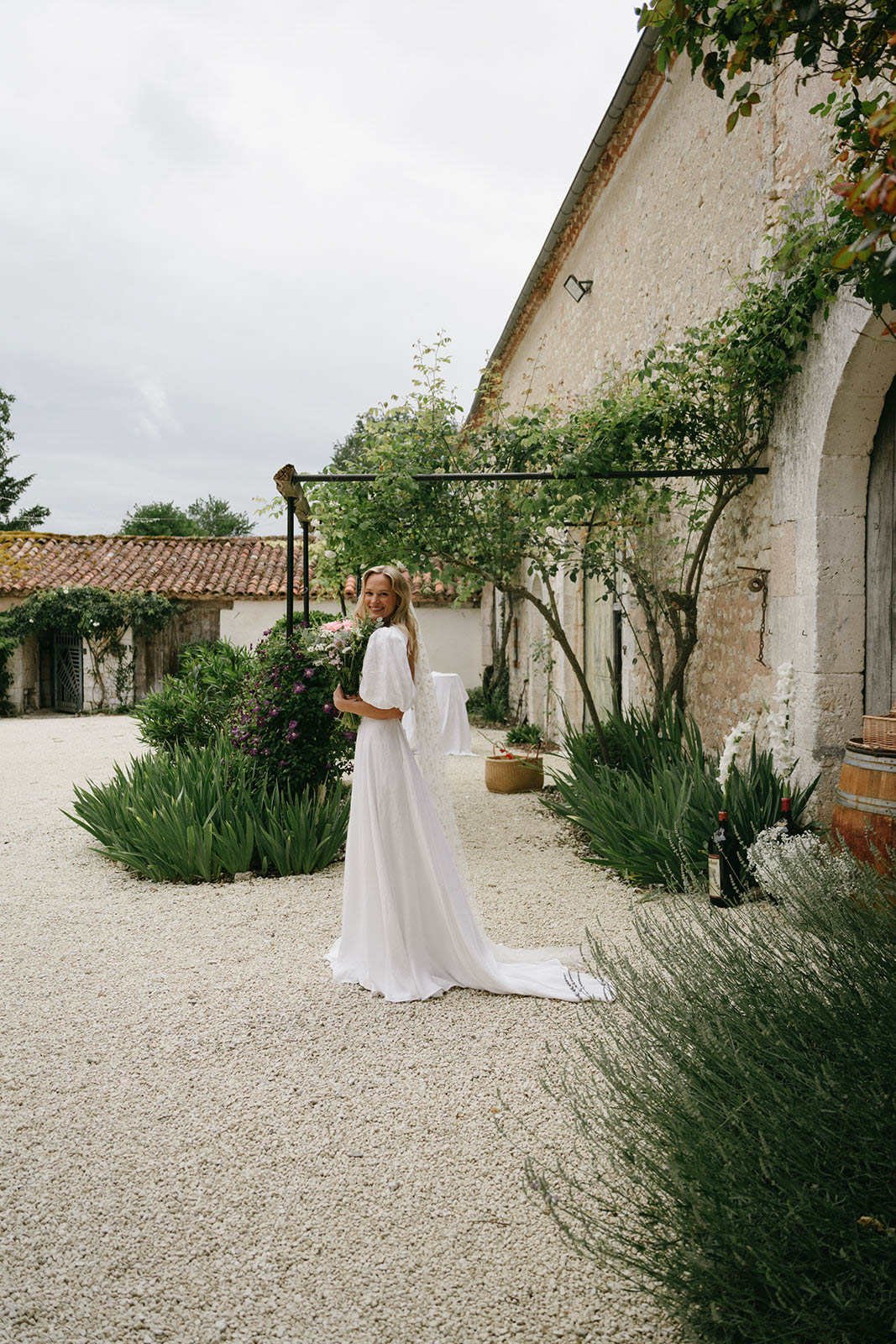 Bride in flowing ivory dress posing in rustic Mediterranean courtyard with stone buildings and lavender plantings