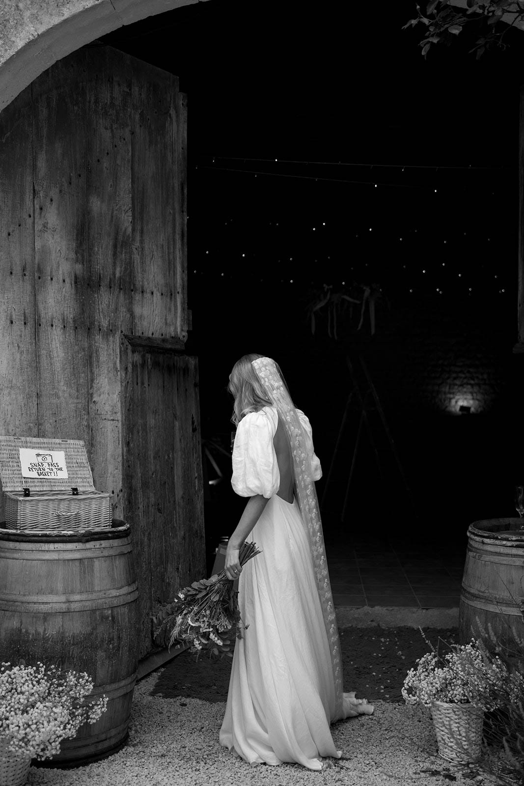 Bride in flowing gown and veil standing in rustic barn doorway looking toward reception guests