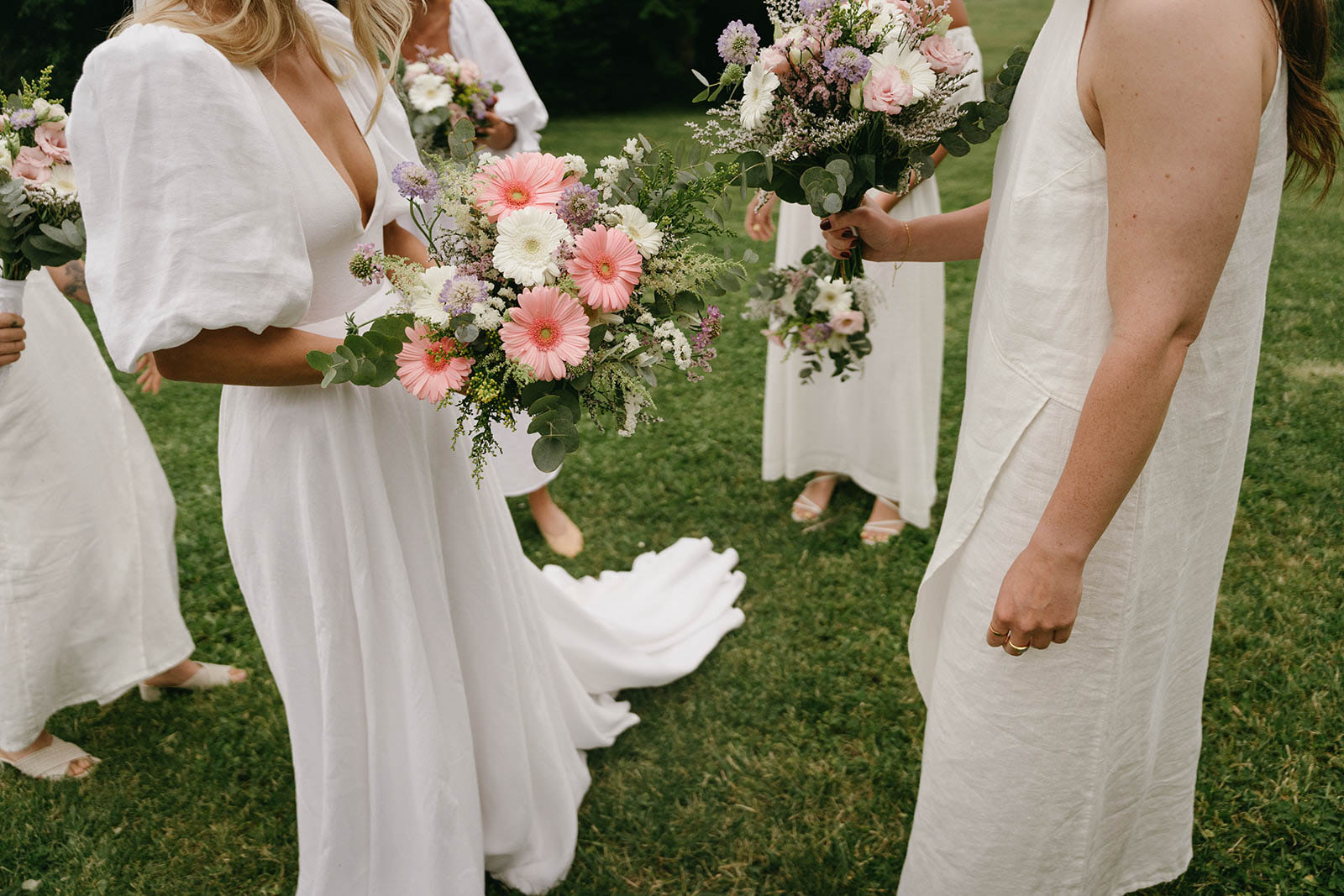 Bride and bridesmaids in ivory linen dresses holding coral and cream bouquet on outdoor lawn