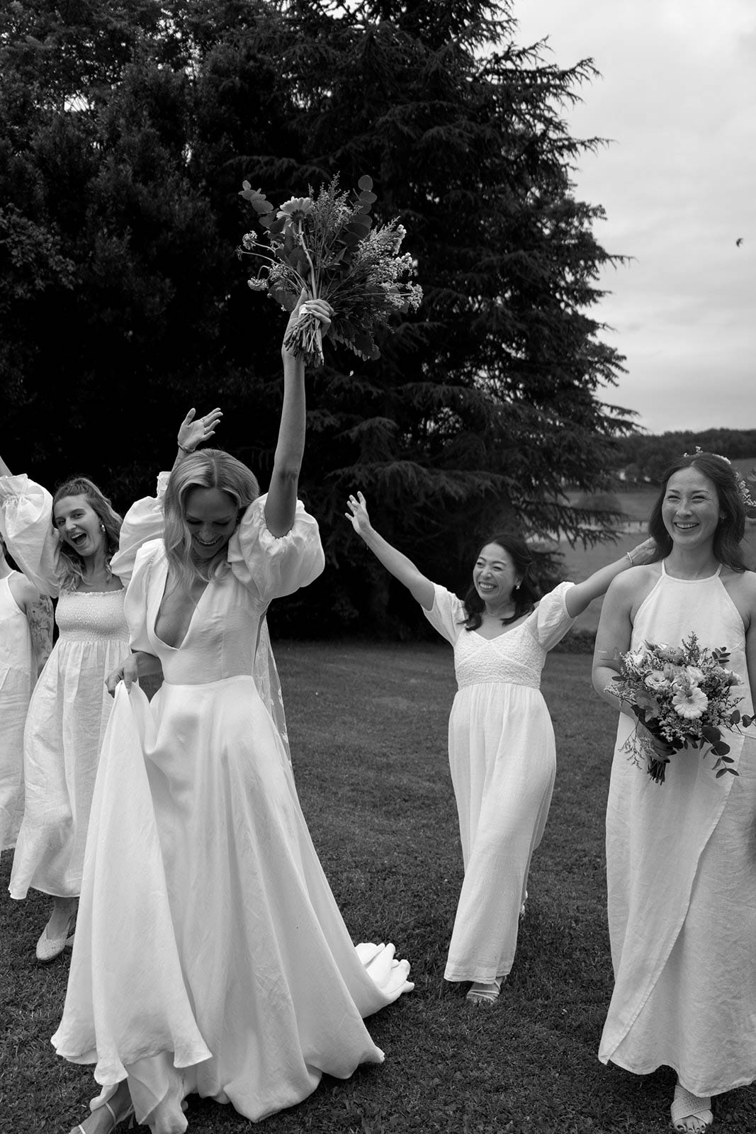 Bride and bridesmaids bouquet toss moment during outdoor wedding reception on lawn with evergreen trees