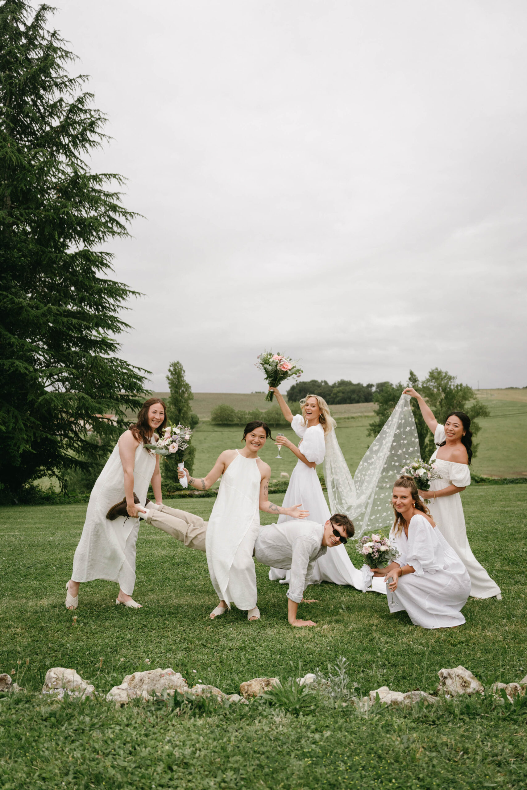 Bride and five bridesmaids in white outfits with pink bouquets in playful candid pose on open lawn
