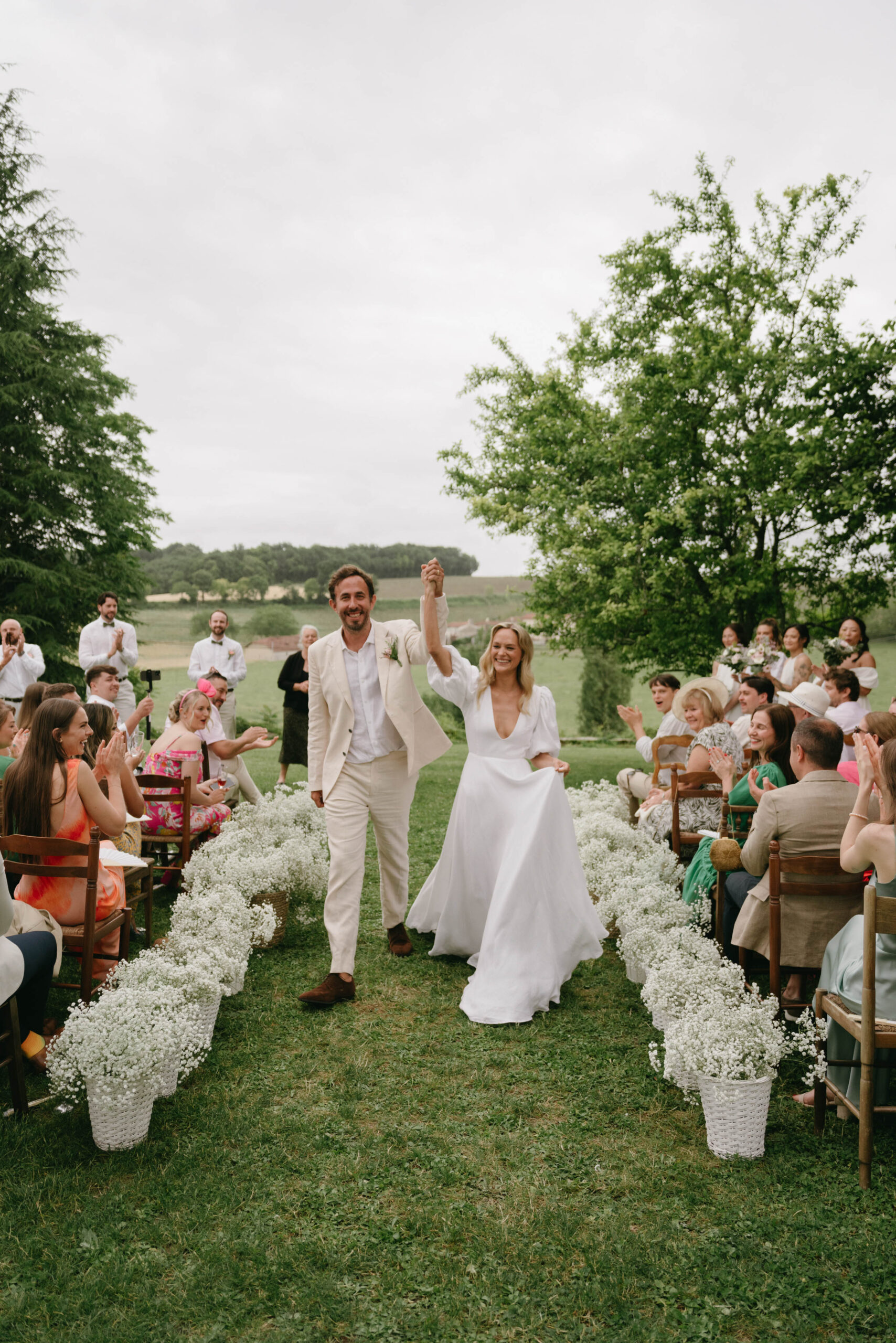 Couple raising joined hands walking back up aisle lined with baby's breath buckets as guests applaud on lawn