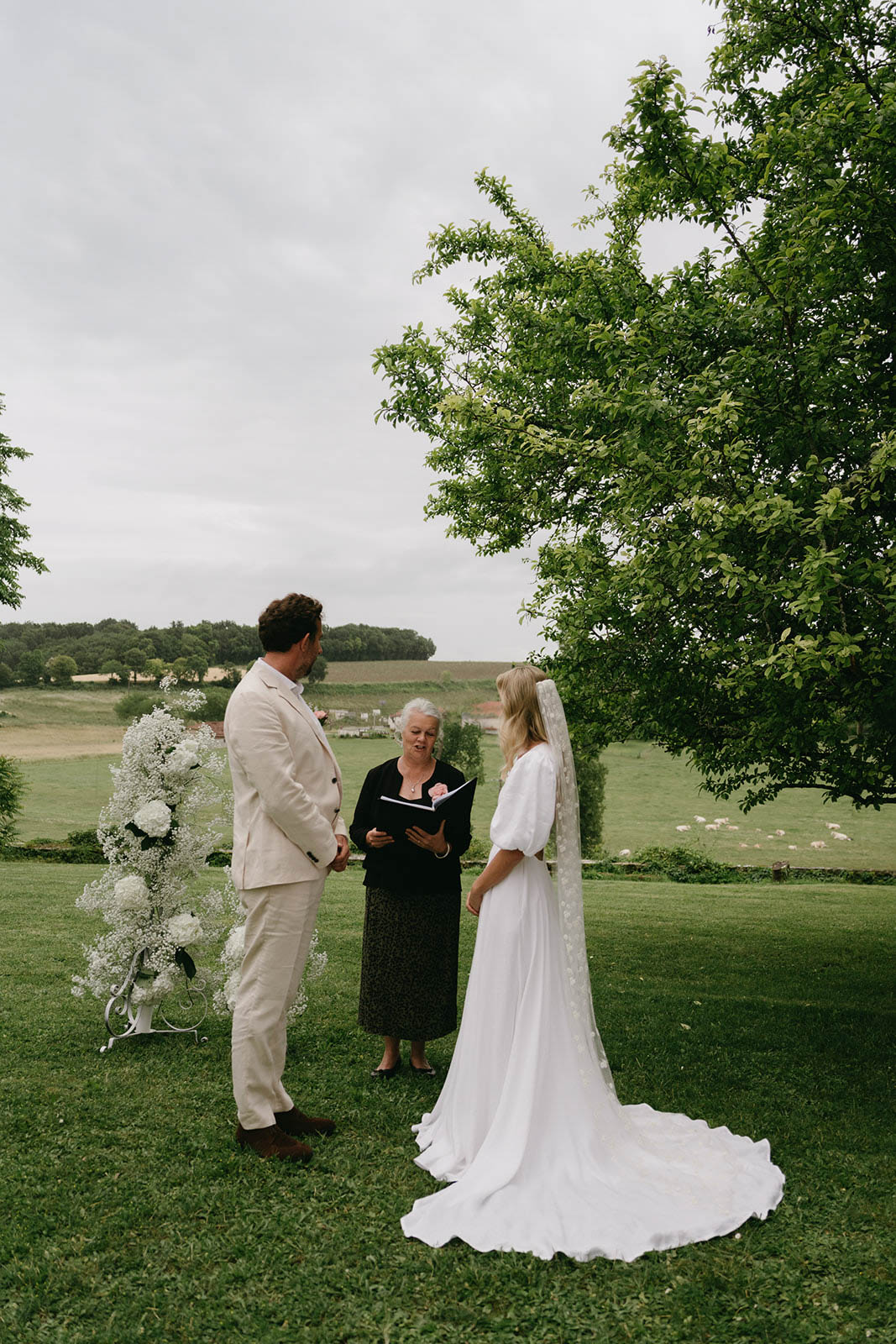 Outdoor wedding ceremony with bride and groom exchanging vows under tree in countryside setting