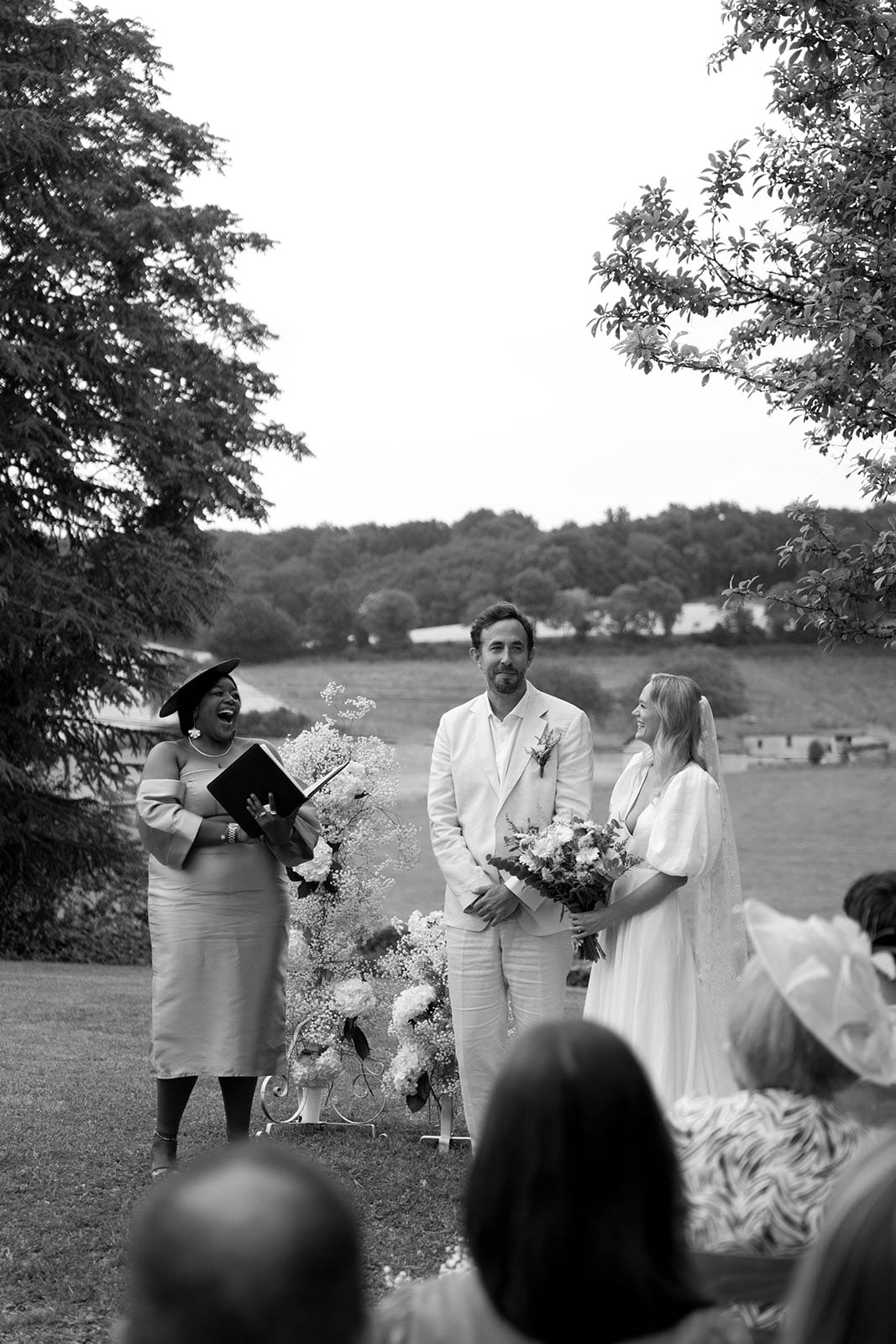 Outdoor wedding ceremony in progress at lakeside venue with couple, officiant, and guests on lawn