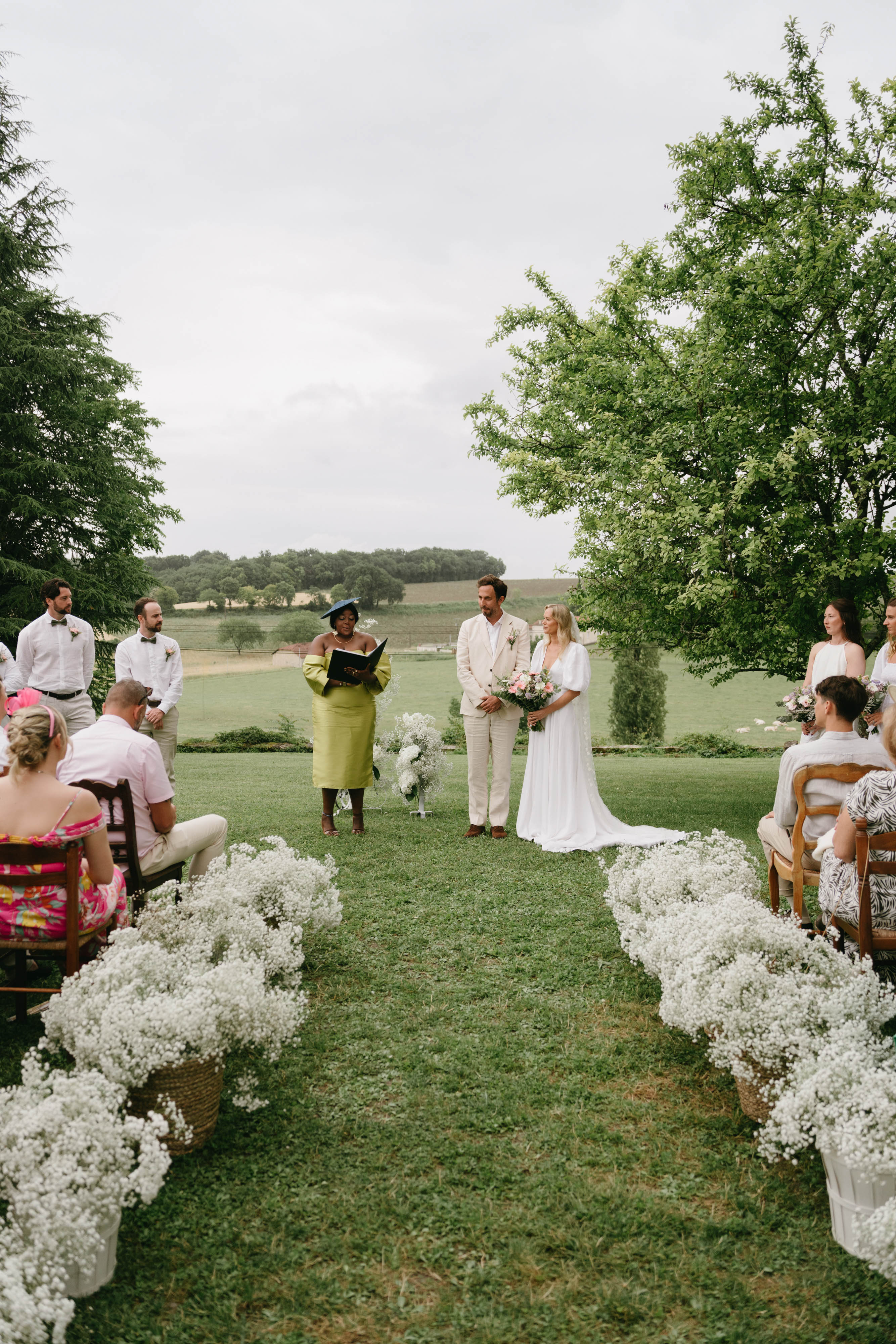 Outdoor wedding ceremony in countryside field with guests seated on wooden chairs and floral aisle arrangements