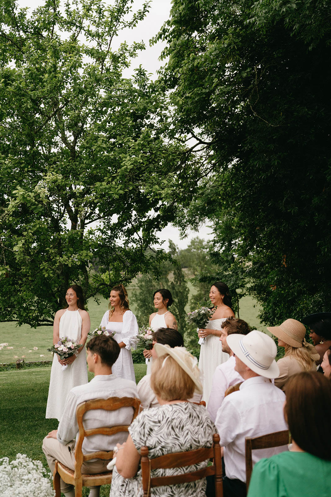 Outdoor wedding ceremony with bridesmaids and seated guests in tree-lined garden setting