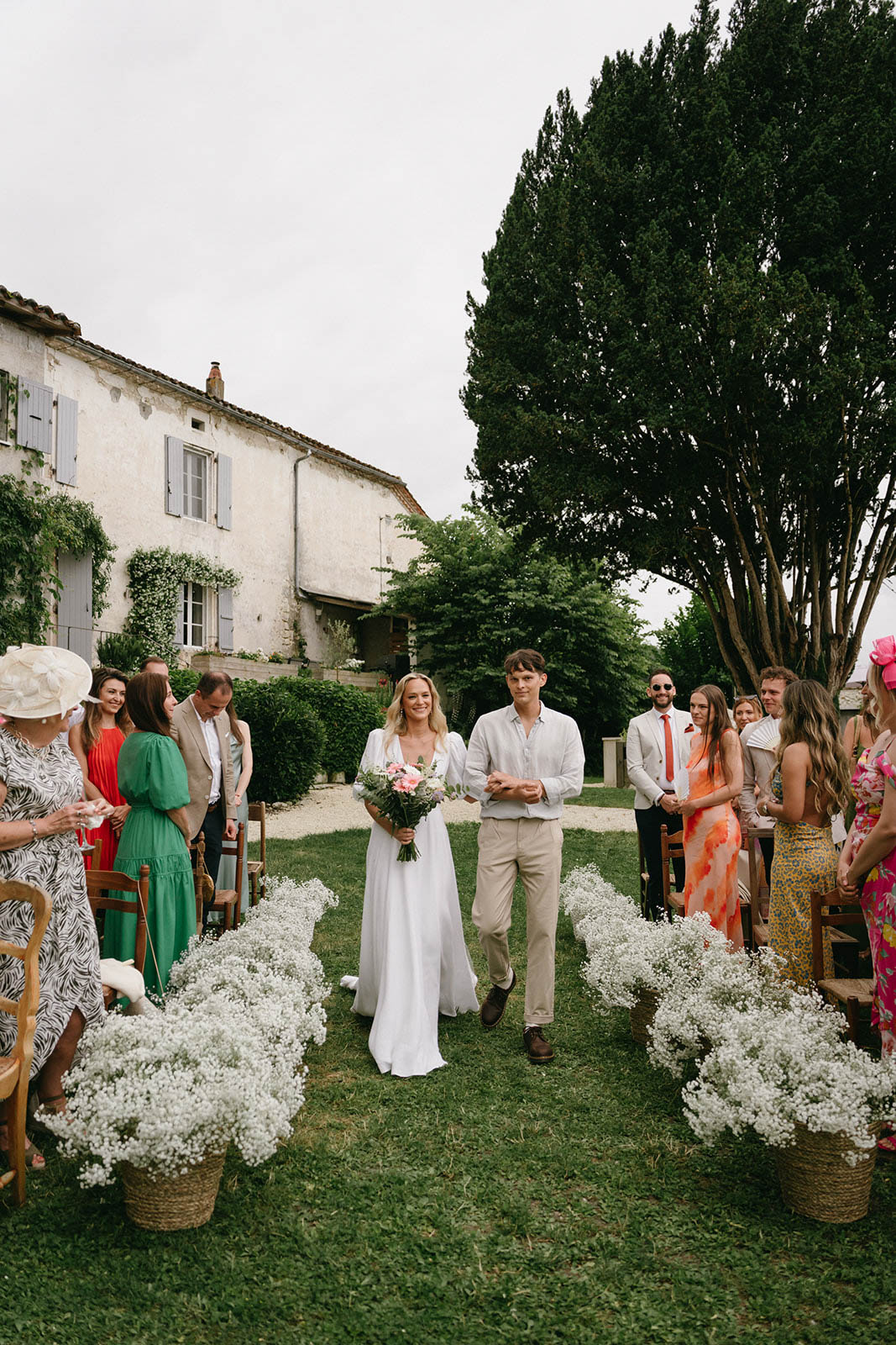Bride and groom walking down outdoor aisle at French countryside wedding ceremony with stone house backdrop