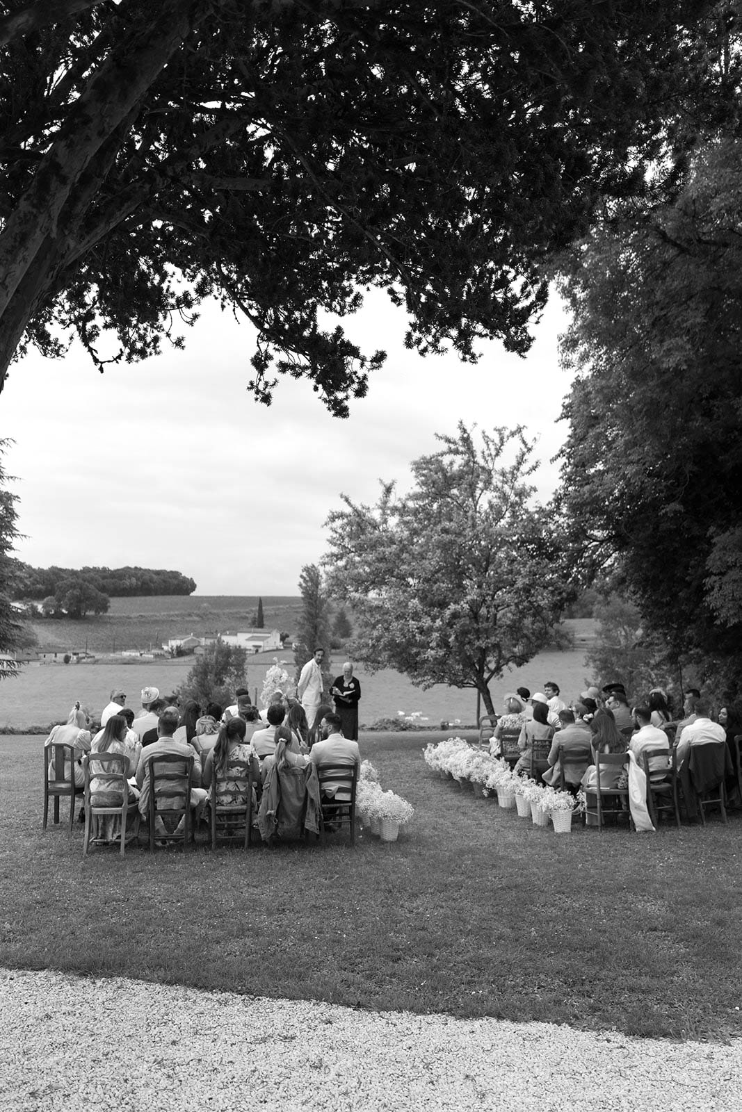 Outdoor wedding ceremony with guests seated in curved rows under trees overlooking countryside landscape