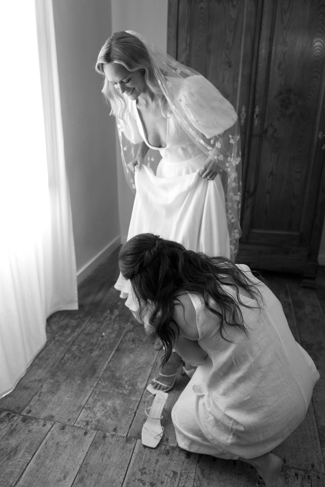 Bride in white slip dress and veil getting shoes adjusted by bridesmaid in indoor bridal preparation moment