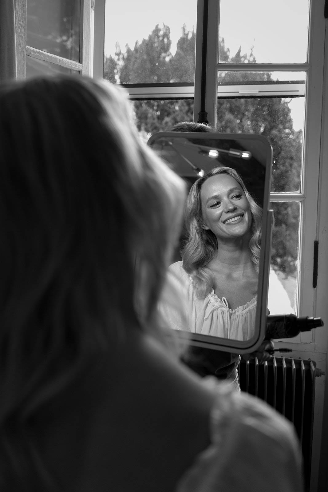 Bride's reflection in mirror during getting ready preparations in interior room with large windows