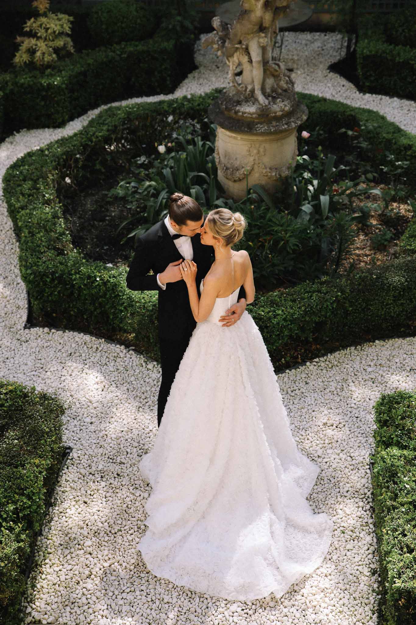 Aerial view of bride and groom kissing in formal parterre garden with geometric hedges and stone urn