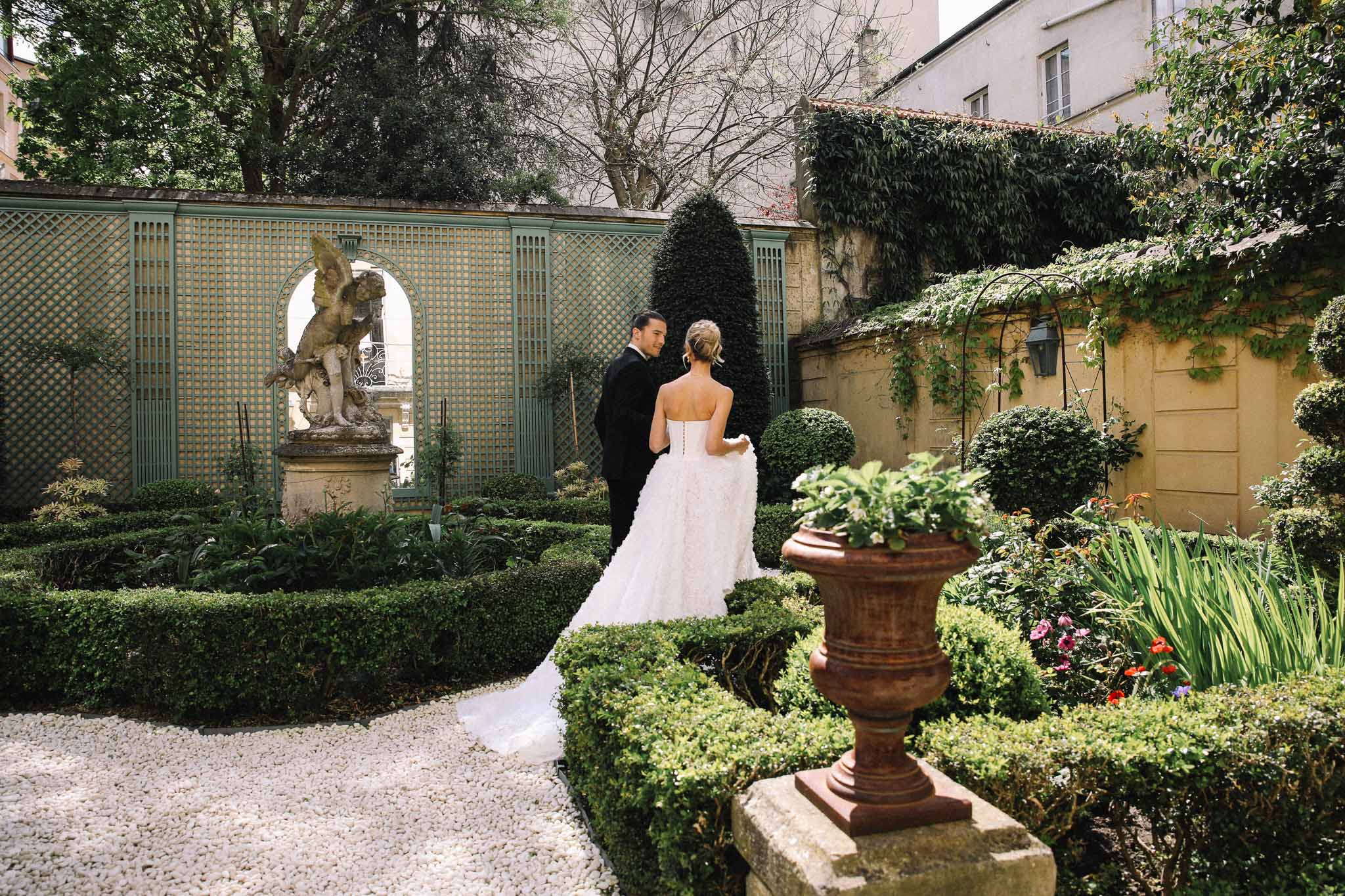 Bride and groom walking through formal French garden courtyard with classical architecture and manicured hedges