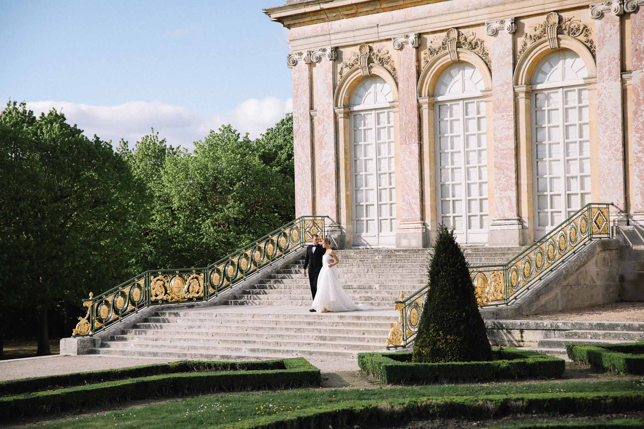 Bride and groom on grand stone staircase of classical chateau with ornate ironwork railings