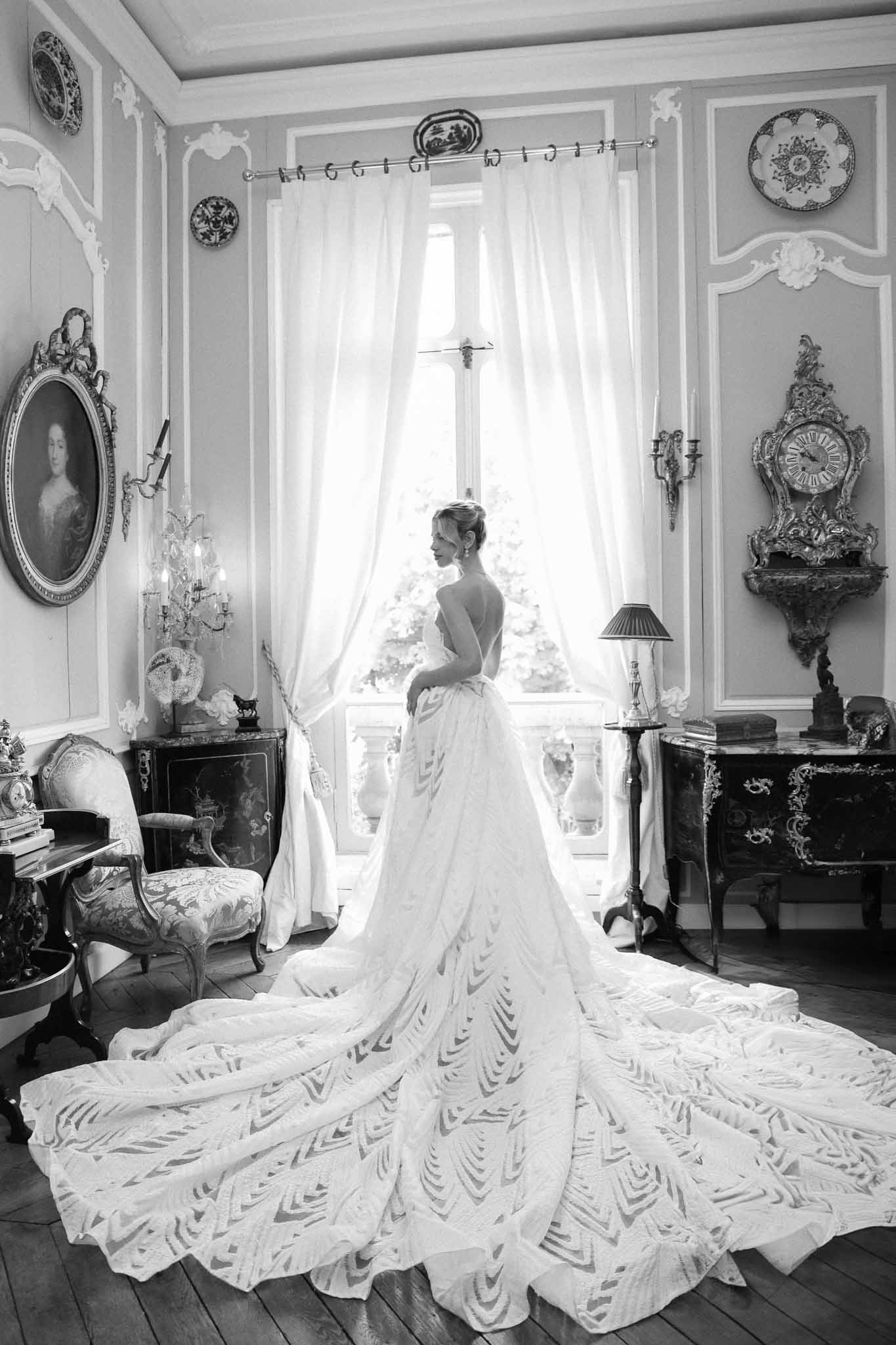 Bride in elaborate gown standing by window in elegant bridal suite with classical European decor