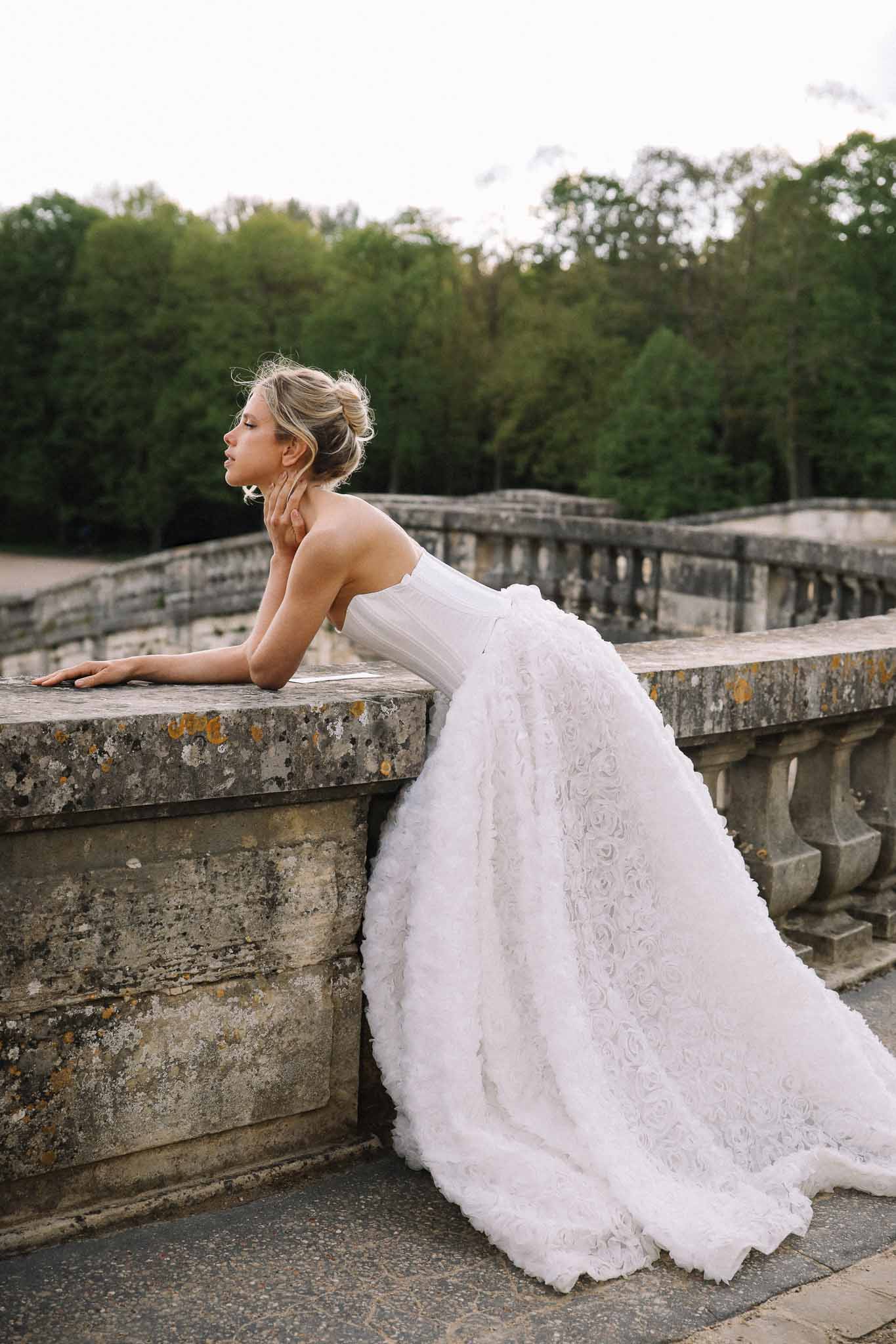 Bride in ivory lace wedding dress posing on historic stone bridge in formal garden setting