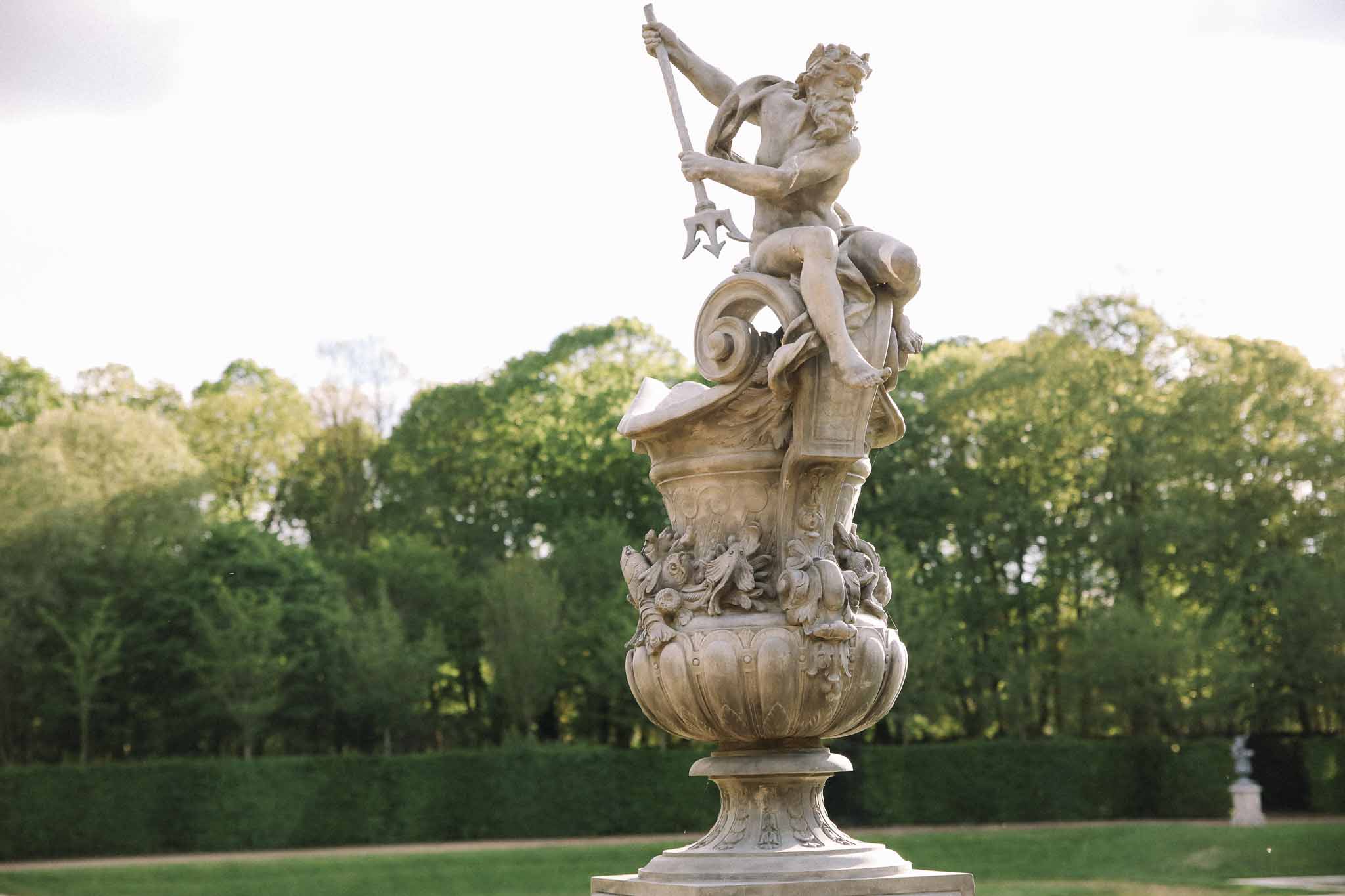 Classical stone cherub sculpture with trident in formal French garden with manicured hedges