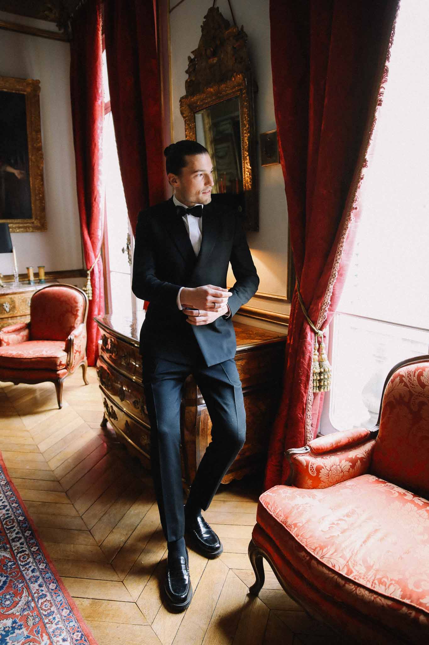 Groom in black tuxedo during getting ready moment in ornate interior room with burgundy curtains