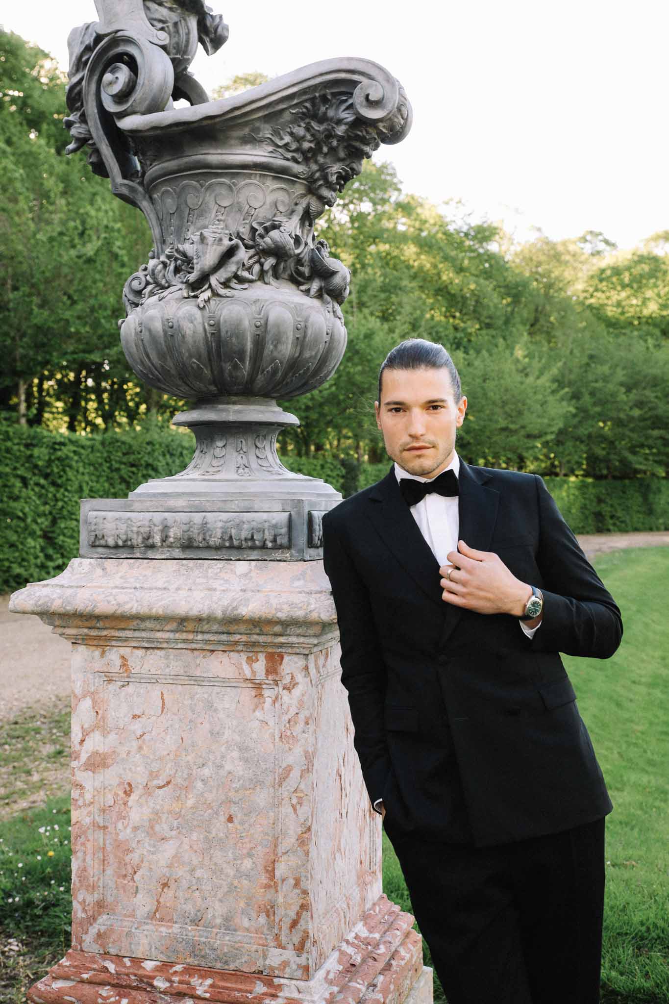 Groom in black tuxedo portrait beside ornamental stone urn in formal garden setting